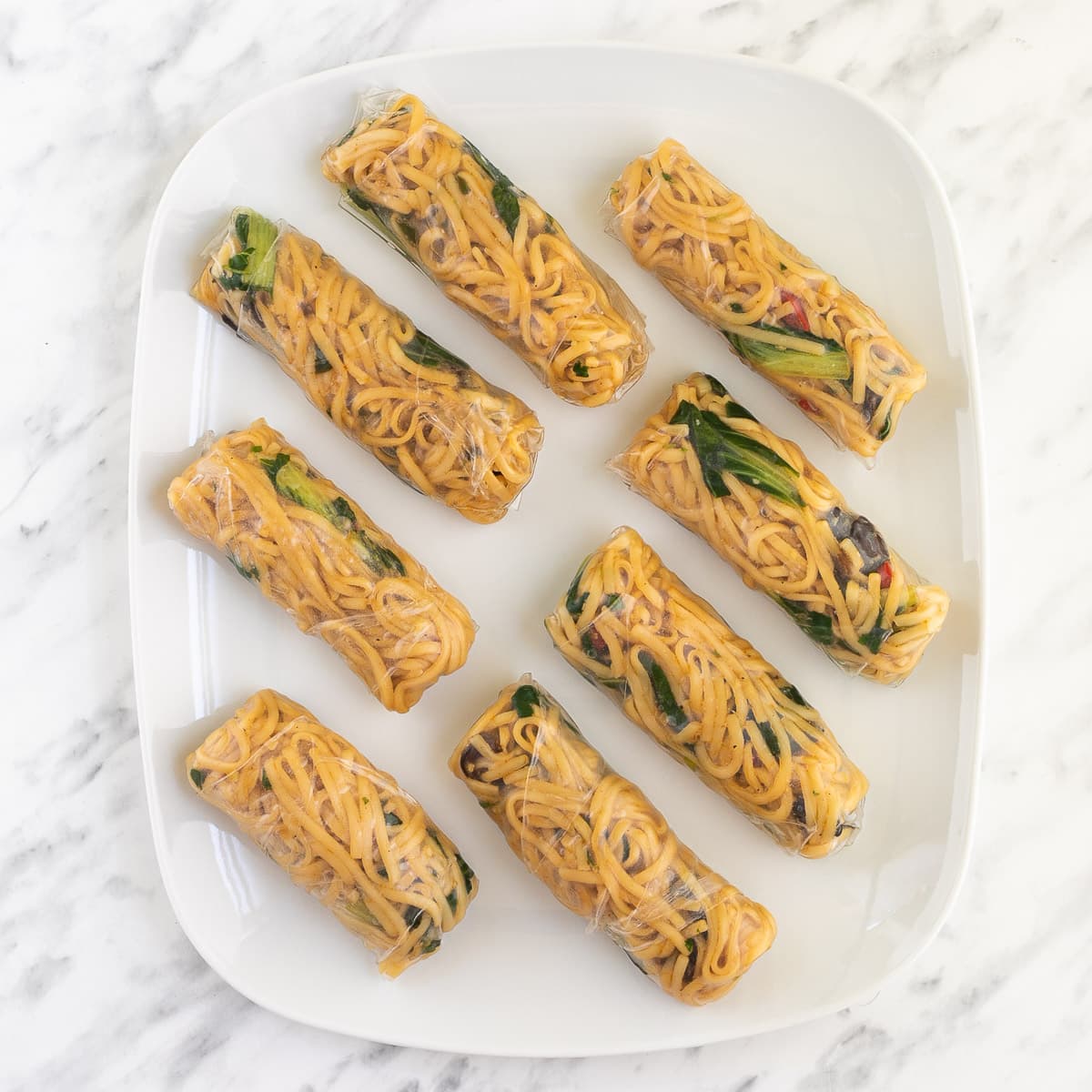 8 ramen spring rolls are placed on a large white plate ready to be fried. Yellow noodles and some green and brown filling is visible.