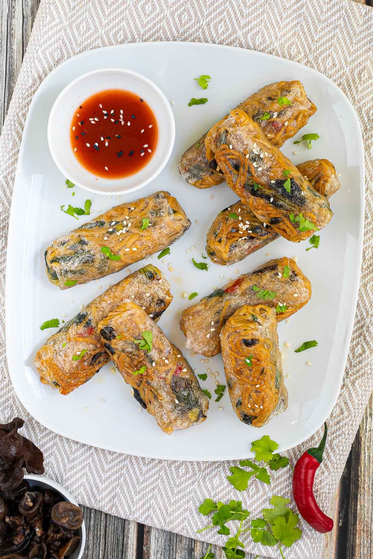 8 ramen spring rolls with yellow ramen noodle filling are placed randomly on a white plate. A red sweet chili sauce is placed in a small white bowl right next to them. They are also sprinkled with chopped green herbs.