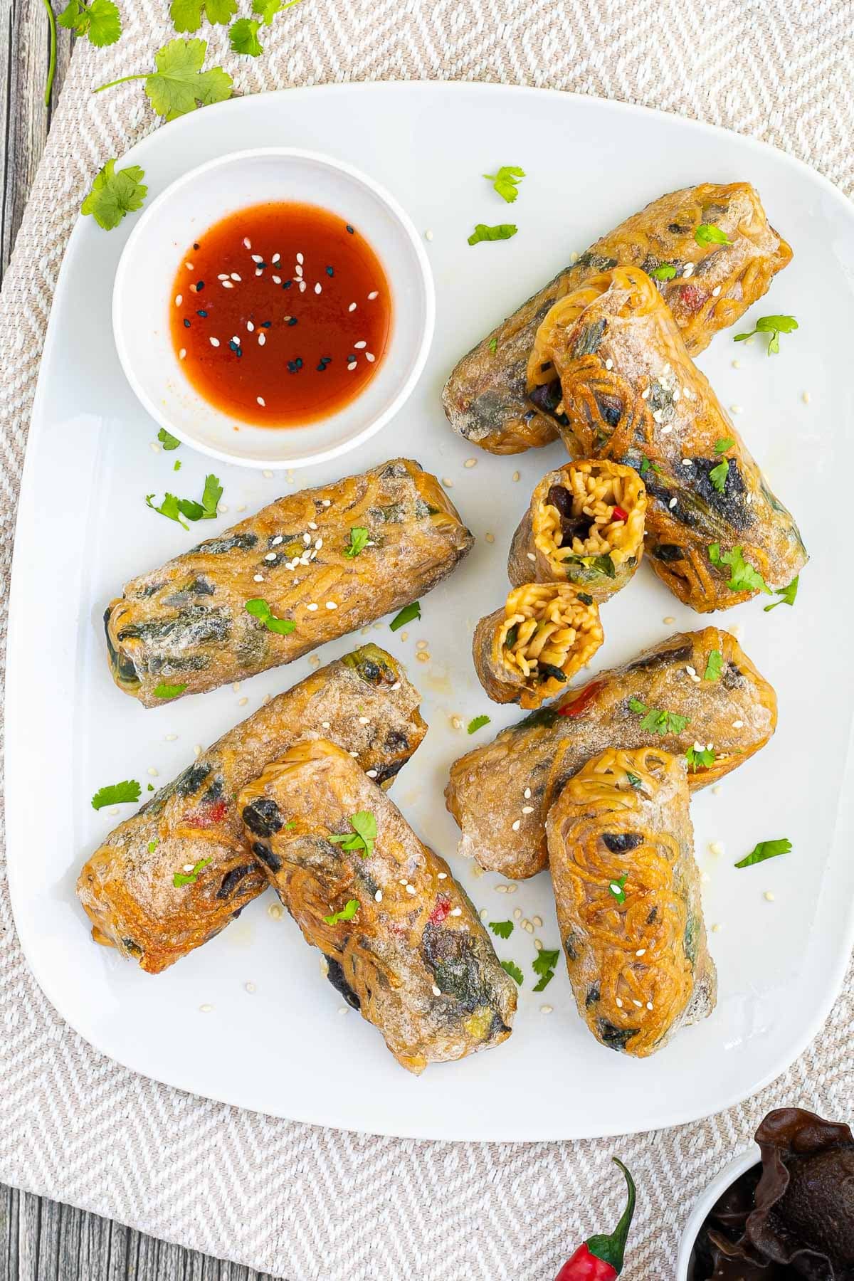 Several ramen spring rolls are on a white plate. A red sweet chili sauce is placed in a small white bowl right next to them. They are also sprinkled with chopped green herbs. 2 rolls are cut in half so the yellow noodles and some green and brown pieces are visible.