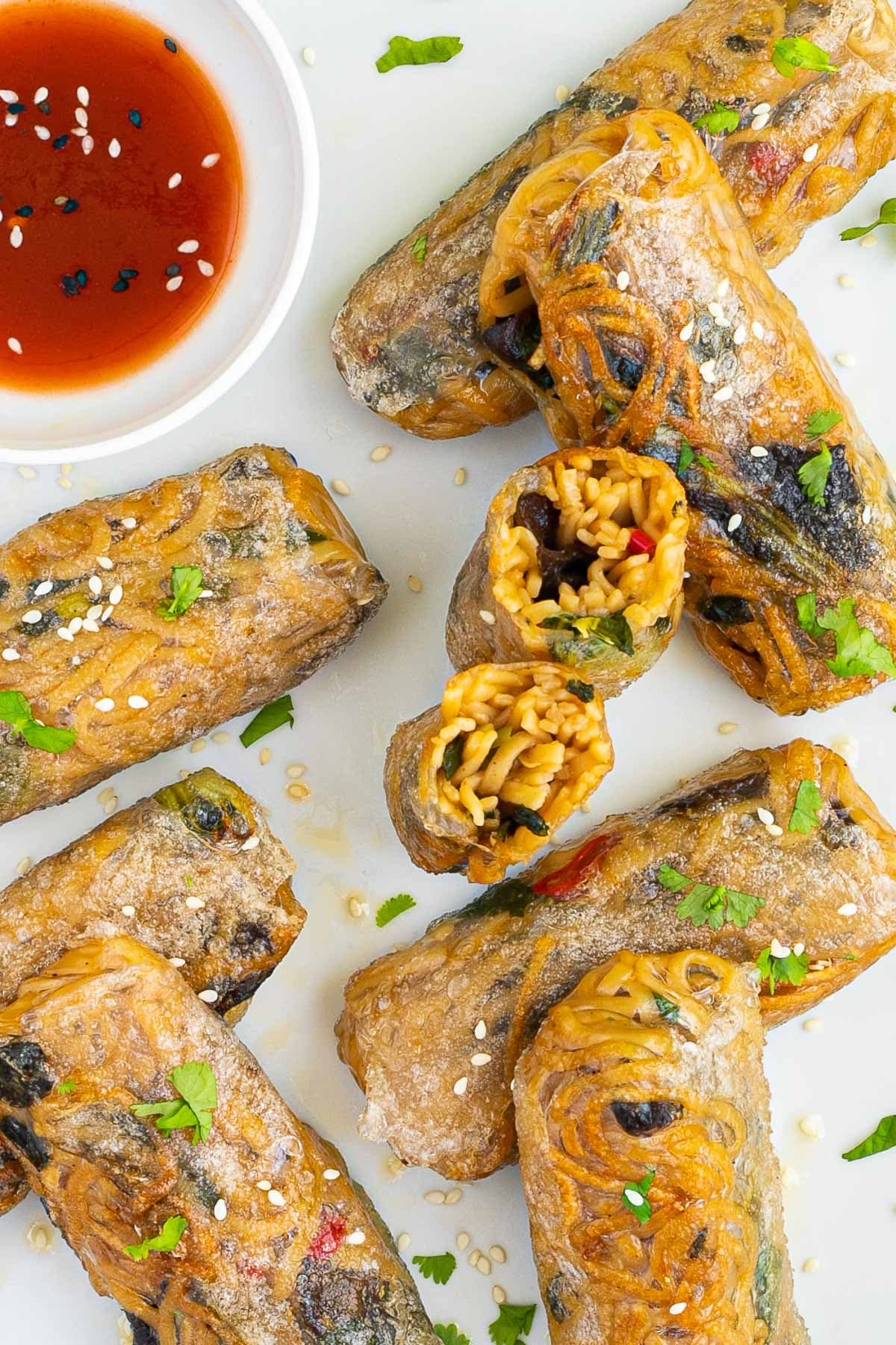 Several ramen spring rolls are on a white plate. A red sweet chili sauce is placed in a small white bowl right next to them. They are also sprinkled with chopped green herbs. 2 rolls are cut in half so the yellow noodles and some green and brown pieces are visible. 