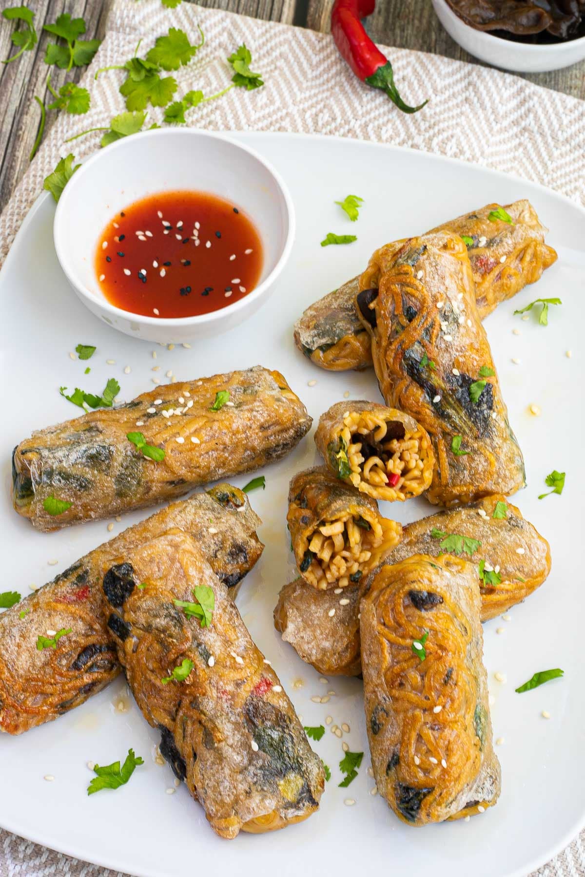 Several ramen spring rolls are on a white plate. A red sweet chili sauce is placed in a small white bowl right next to them. They are also sprinkled with chopped green herbs. 2 rolls are cut in half so the yellow noodles and some green and brown pieces are visible.