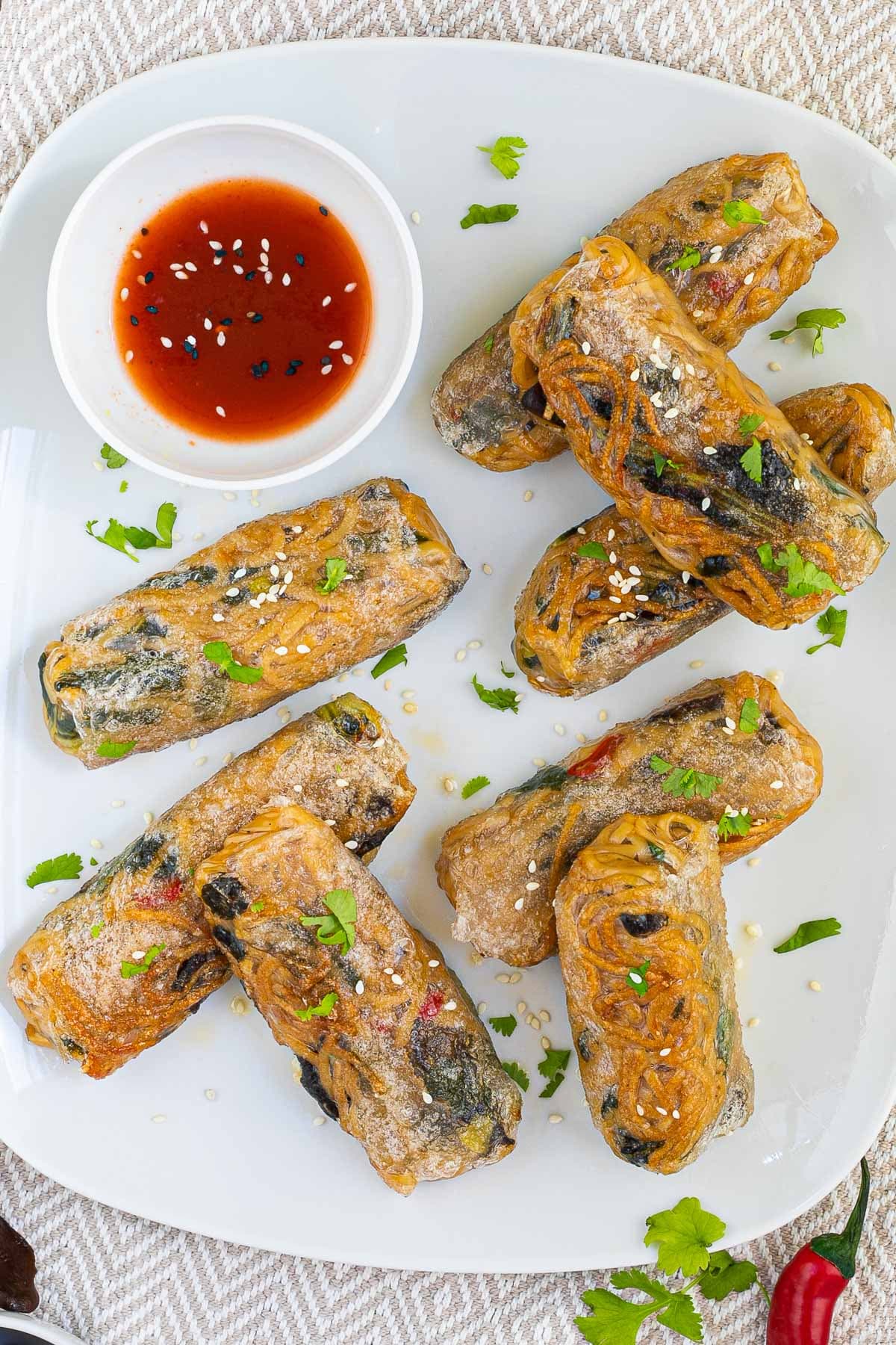 8 ramen spring rolls with yellow ramen noodle filling are placed randomly on a white plate. A red sweet chili sauce is placed in a small white bowl right next to them. They are also sprinkled with chopped green herbs.