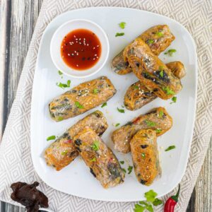 8 ramen spring rolls with yellow ramen noodle filling are placed randomly on a white plate. A red sweet chili sauce is placed in a small white bowl right next to them. They are also sprinkled with chopped green herbs.