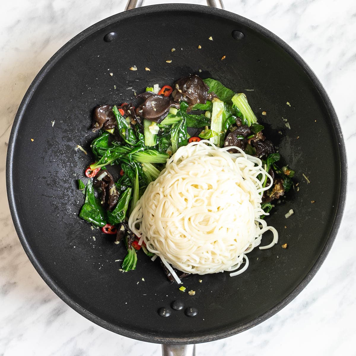 Wok with wilted chopped bok choy, crispy mushroom slices, red chili pepper slices, and chopped onion topped with a heap of ramen noodles.