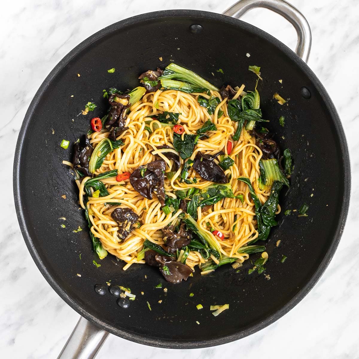 Wok with wilted chopped bok choy, crispy mushroom slices, red chili pepper slices, and yellow ramen noodles mixed.