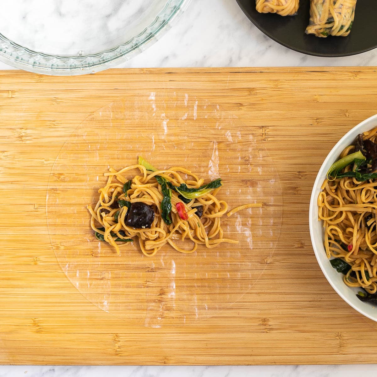 A wooden cutting board with wet rice paper topped with ramen noodles in the middle. Ramen noodles in a white bowl. Water in a glass bowl and 2 finish spring rolls on a black plate around it.