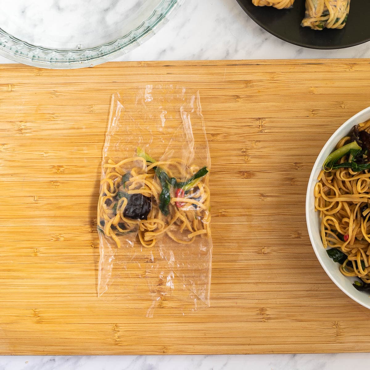 A wooden cutting board with wet rice paper with ramen noodle filling folded in the middle from the sides. Ramen noodles in a white bowl. Water in a glass bowl and 2 finish spring rolls on a black plate around it.