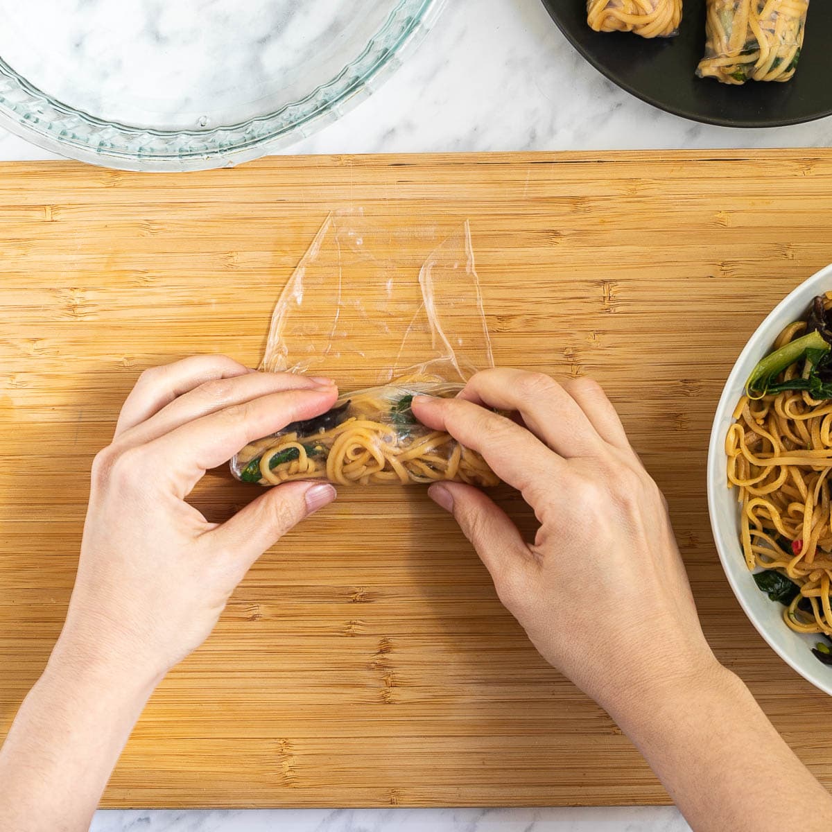 A hand is rolling a rice paper with the filling inside from bottom to top. Ramen noodles in a white bowl. Water in a glass bowl and 2 finish spring rolls on a black plate around it.