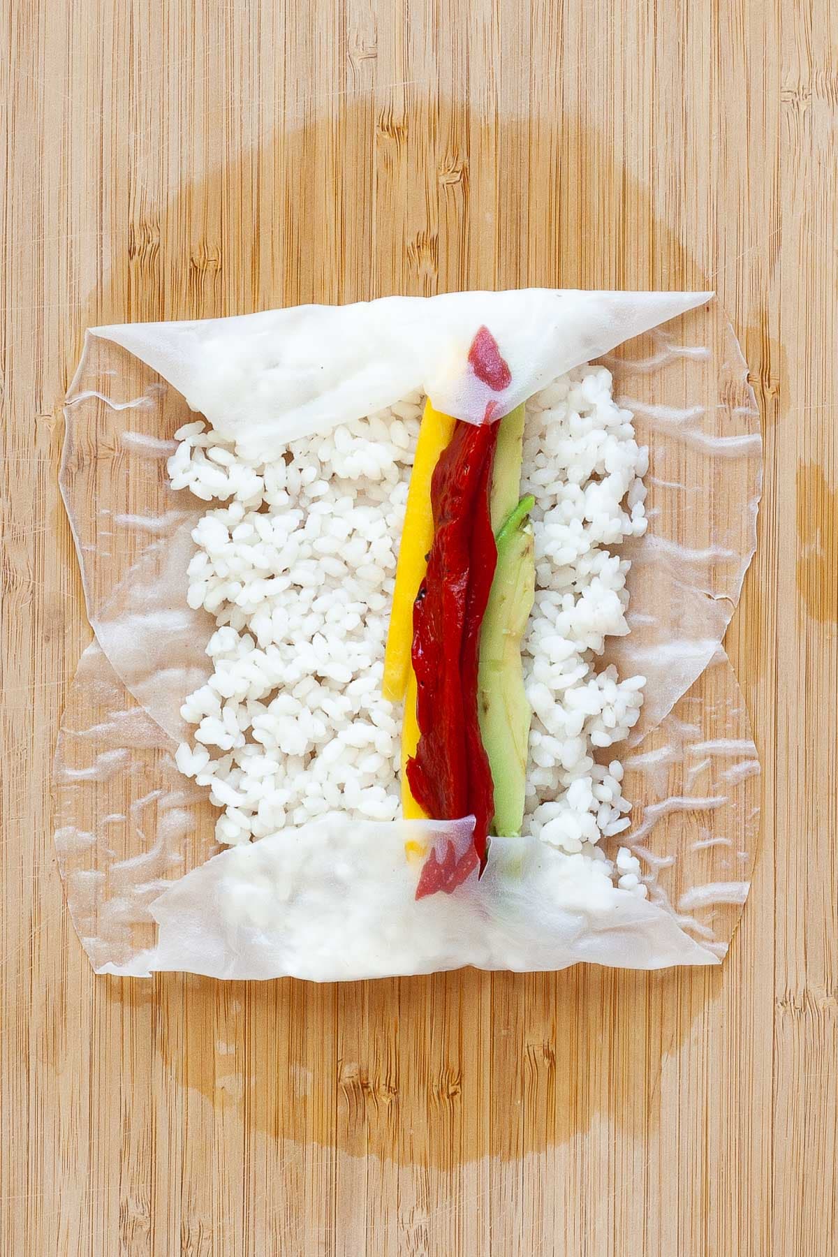 Soft wet rice paper sheets with rice, red pepper, mango, and avocado slices on top on a wooden board