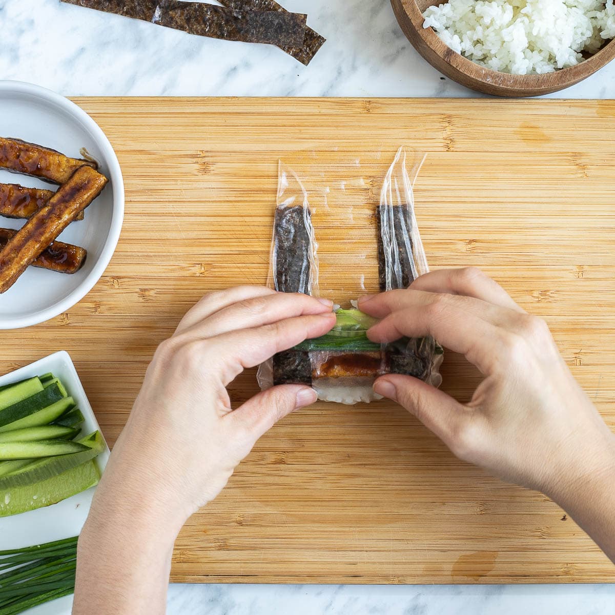 A hand is rolling a rice paper with the filling inside from bottom to top. The leftover fillings are in small bowls like rice, cucumber sticks, avocado slices, chives, brown sticky tofu steaks and nori strips.