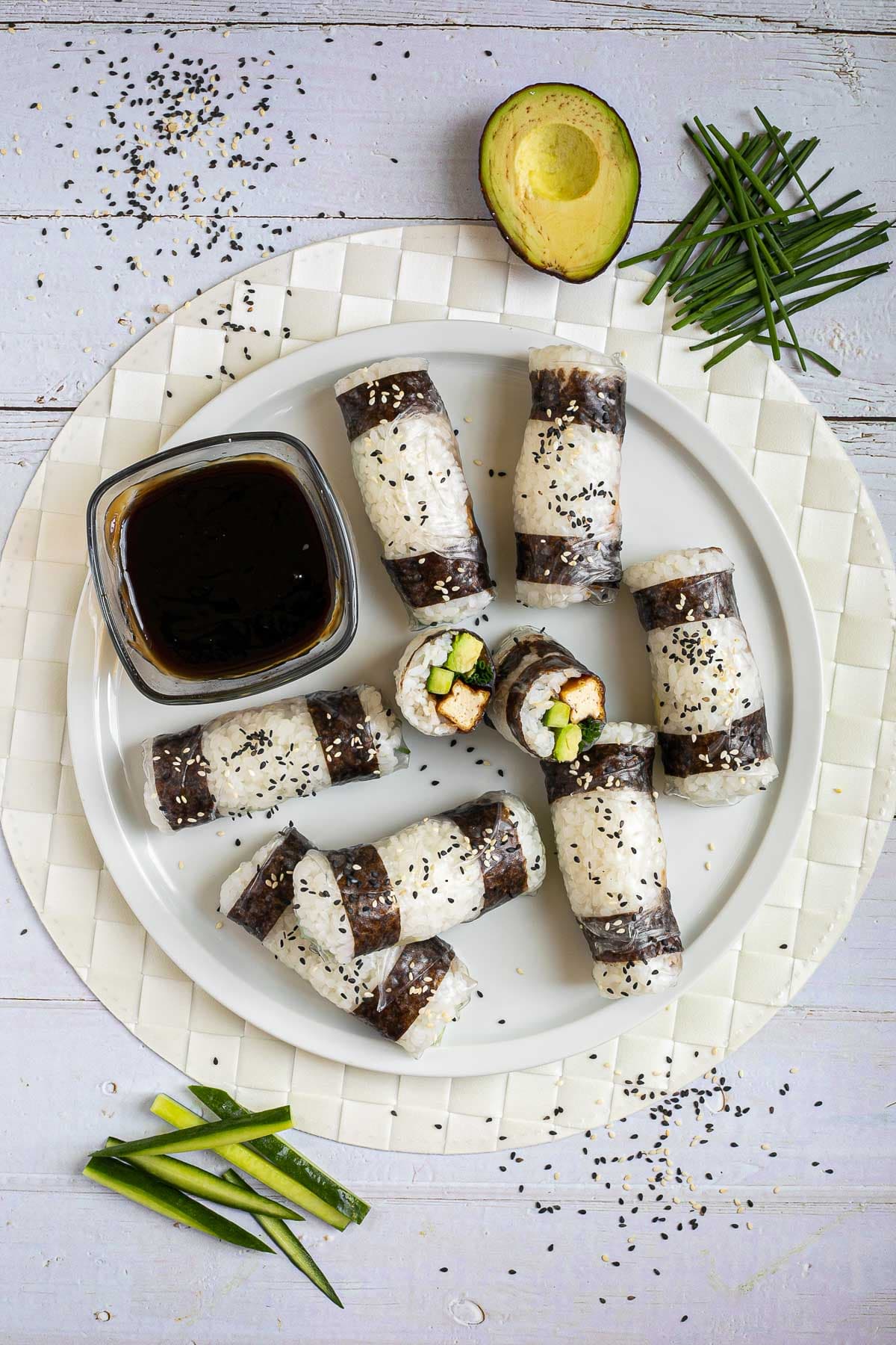 Black and white rice paper rolls on a large white plate sprinkled with black and white sesame seeds. One is cut in the middle so the filling of tofu sticks, cucumber, avocado, and chives is visible.