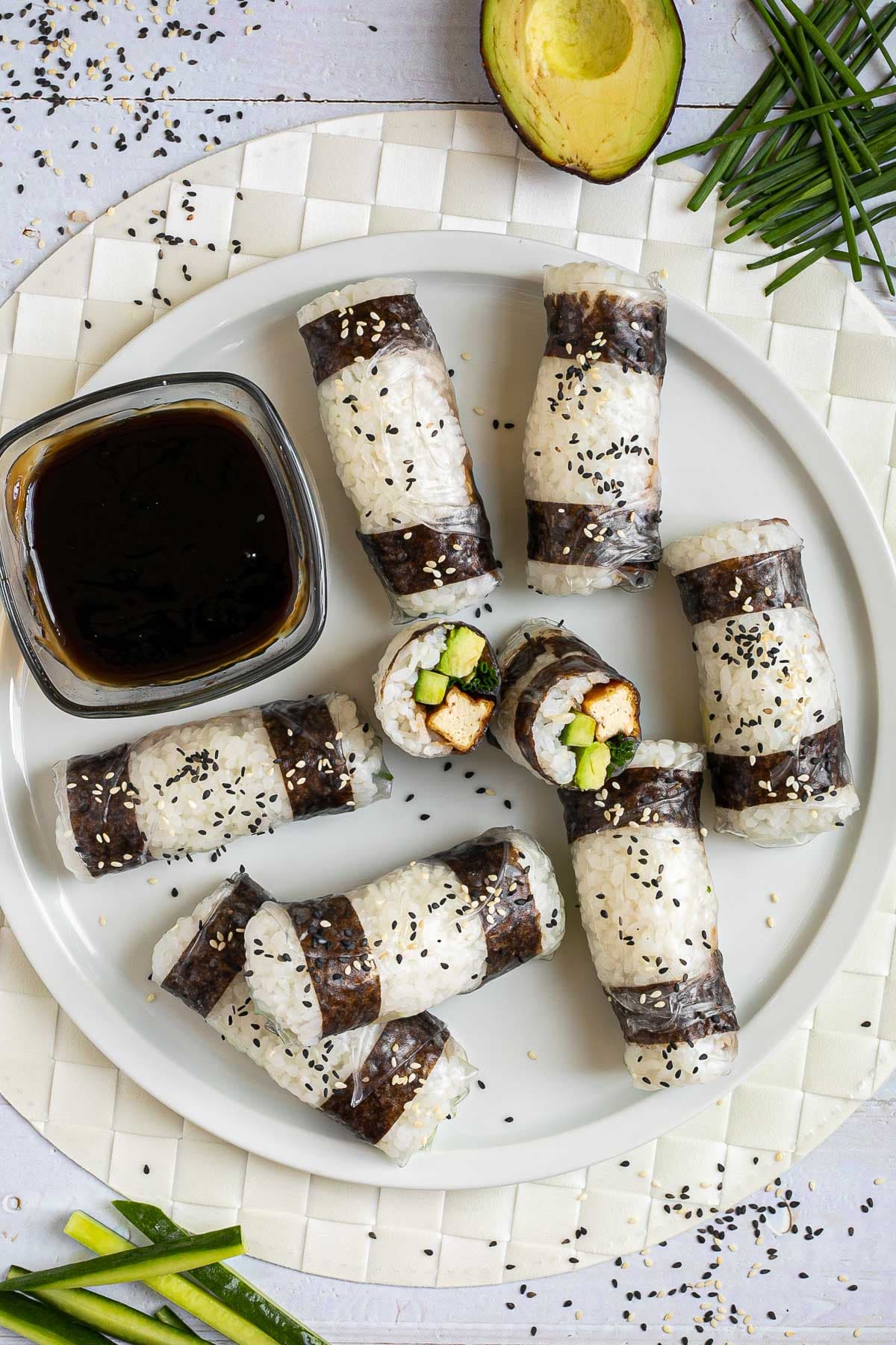 Black and white rice paper rolls on a large white plate sprinkled with black and white sesame seeds. One is cut in the middle so the filling of tofu sticks, cucumber, avocado, and chives is visible.