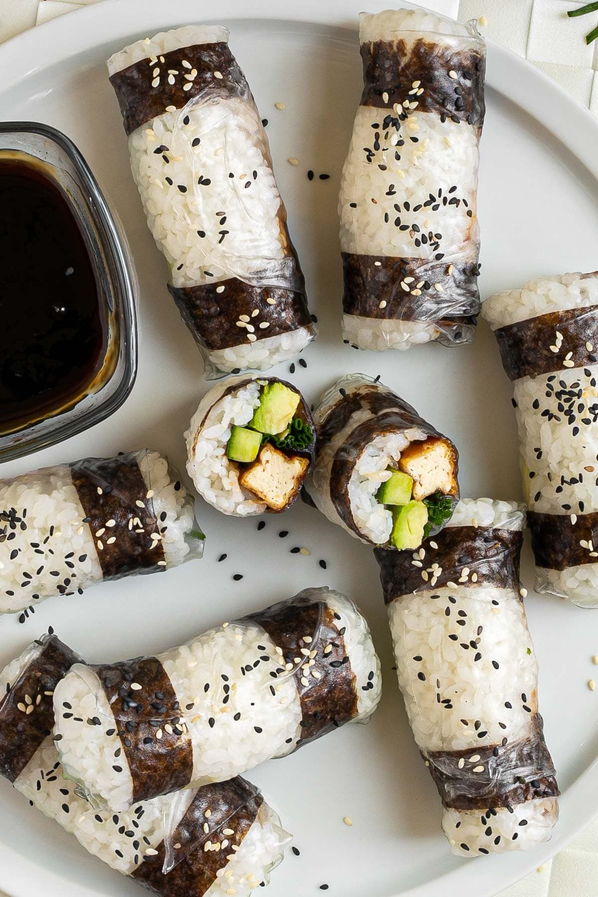 Black and white rice paper rolls on a large white plate sprinkled with black and white sesame seeds. One is cut in the middle so the filling of tofu sticks, cucumber, avocado, and chives is visible.