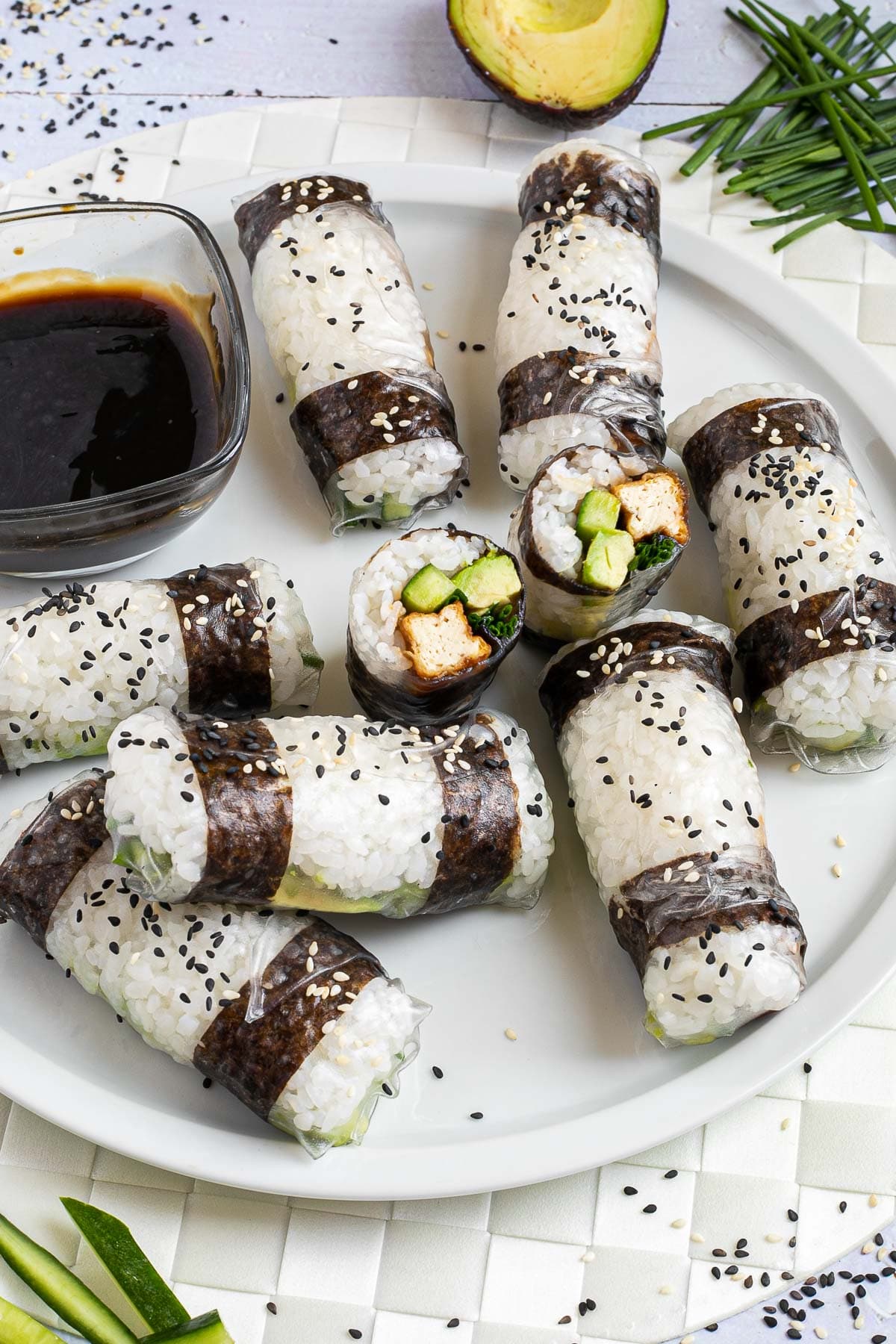 Black and white rice paper rolls on a large white plate sprinkled with black and white sesame seeds. One is cut in the middle so the filling of tofu sticks, cucumber, avocado, and chives is visible.