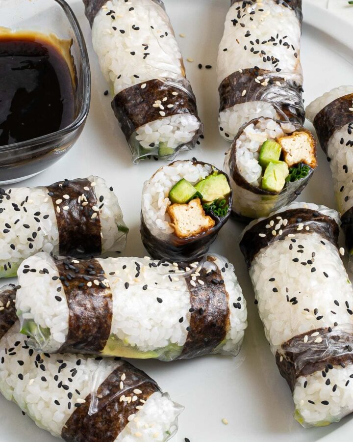 Black and white rice paper rolls on a large white plate sprinkled with black and white sesame seeds. One is cut in the middle so the filling of tofu sticks, cucumber, avocado, and chives is visible.