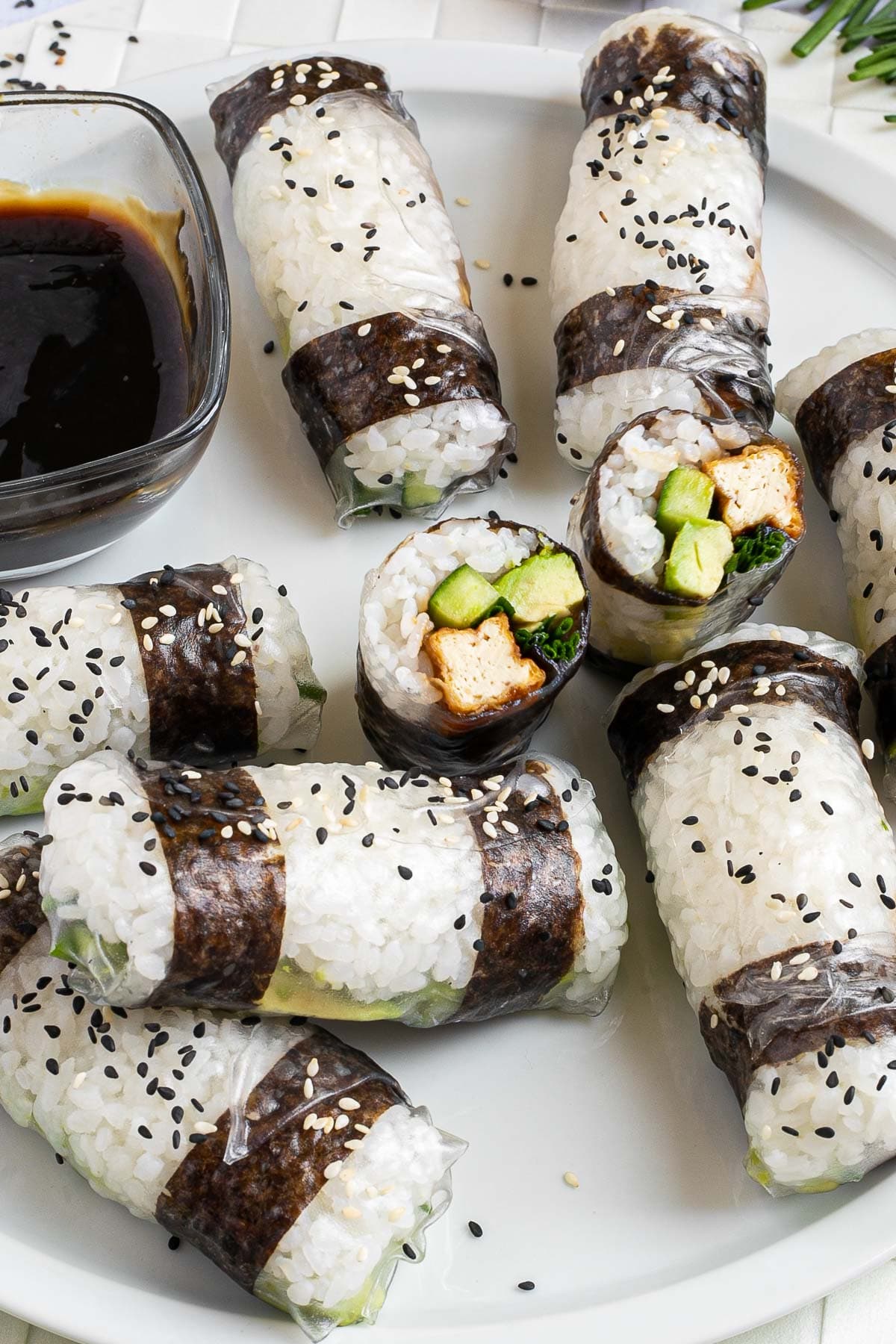 Black and white rice paper rolls on a large white plate sprinkled with black and white sesame seeds. One is cut in the middle so the filling of tofu sticks, cucumber, avocado, and chives is visible.