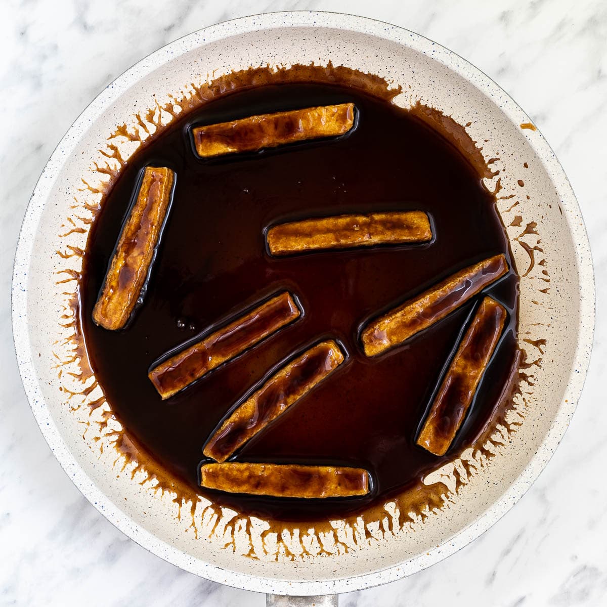 A white frying pan with tofu sticks in a dark brown liquid.