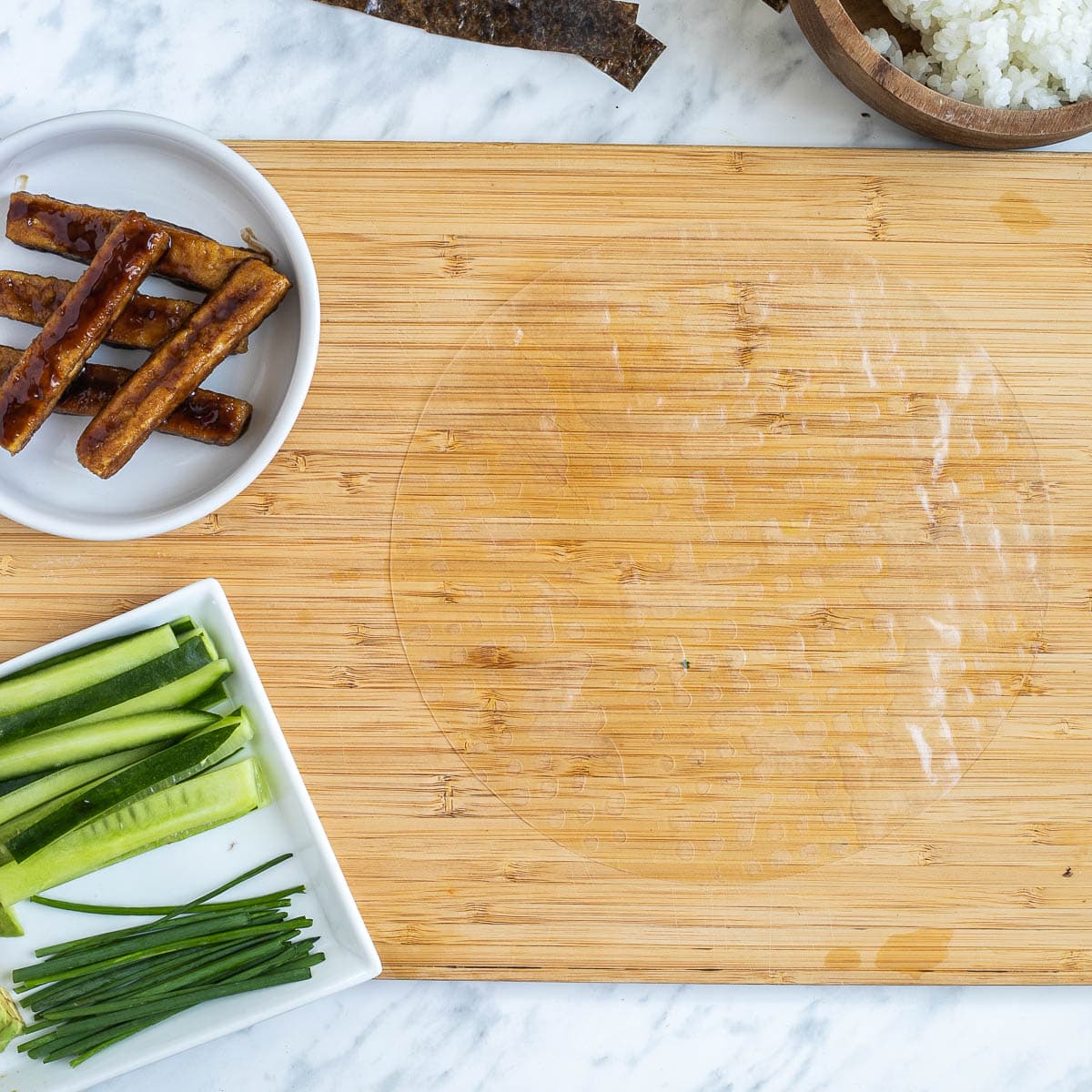 A wooden cutting board with wet rice paper. The fillings are in small bowls like rice, cucumber sticks, avocado slices, chives, brown sticky tofu steaks and nori strips.