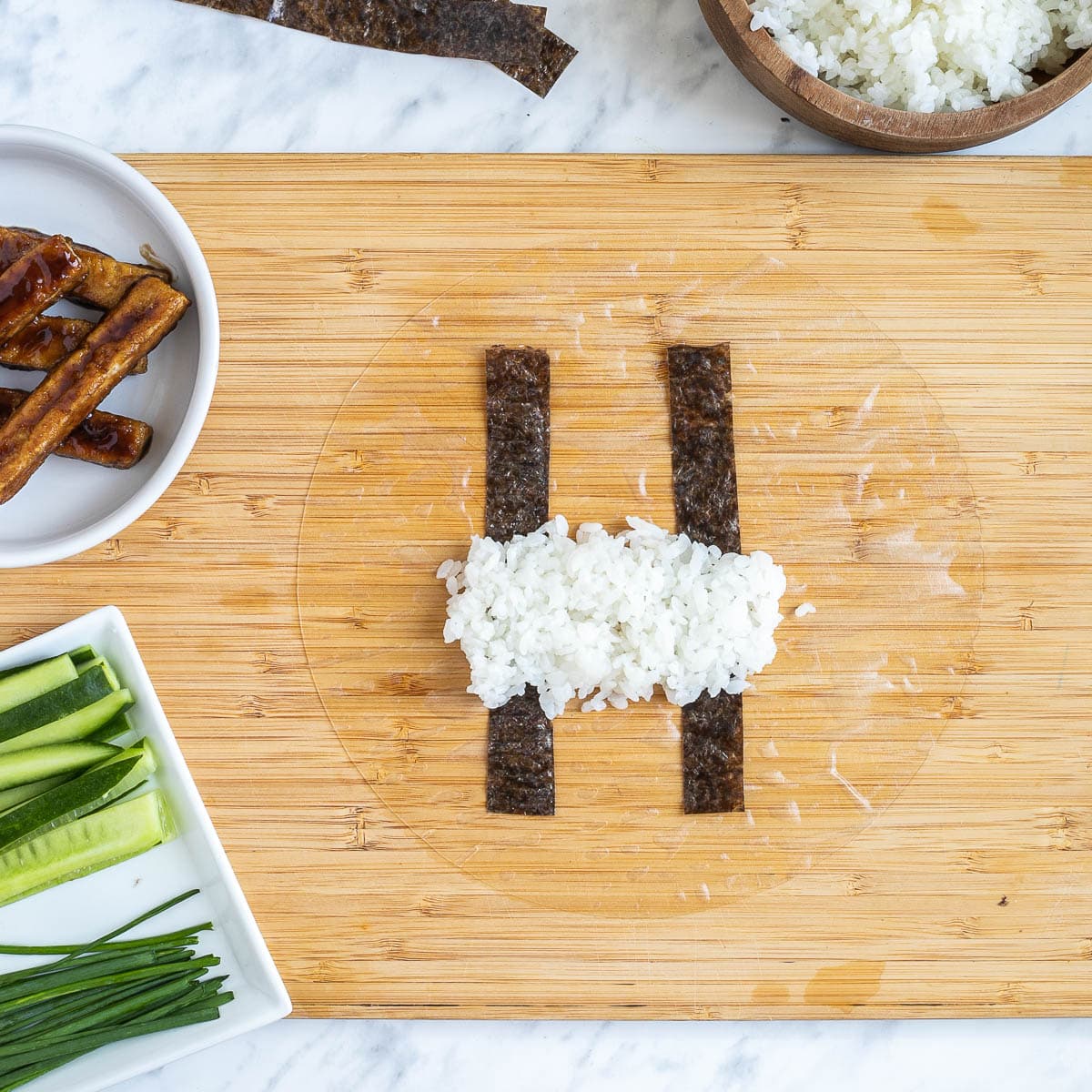 A wooden cutting board with wet rice paper with two thin nori strips and rice in the middle. The fillings are in small bowls like rice, cucumber sticks, avocado slices, chives, brown sticky tofu steaks and nori strips.