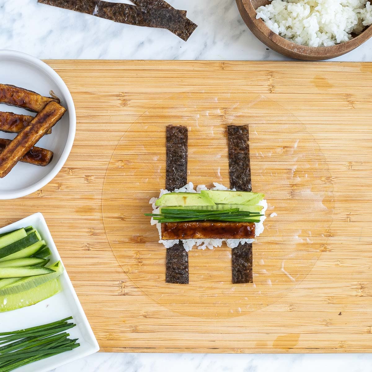 A wooden cutting board with wet rice paper, two nori strips, rice, cucumber sticks, avocado slices, chives, and brown tofu sticks in the middle. The remaining fillings are in small bowls.