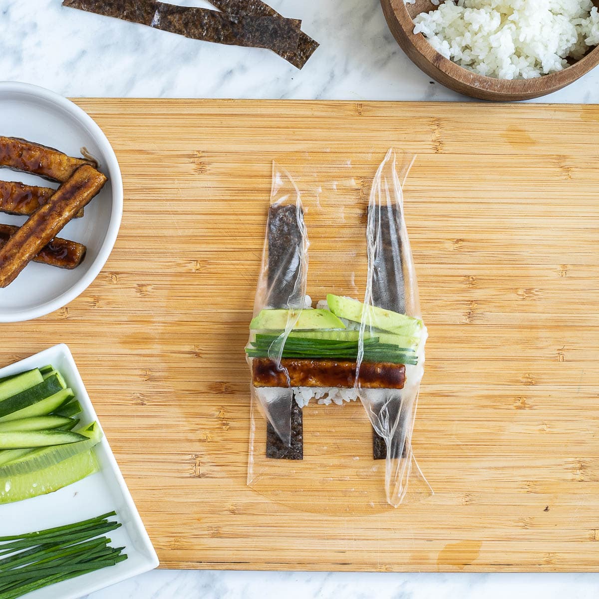 A wooden cutting board with wet rice paper folded in the middle from the sides with two nori strips, rice, cucumber sticks, avocado slices, chives, and brown tofu sticks in the middle. The remaining fillings are in small bowls.