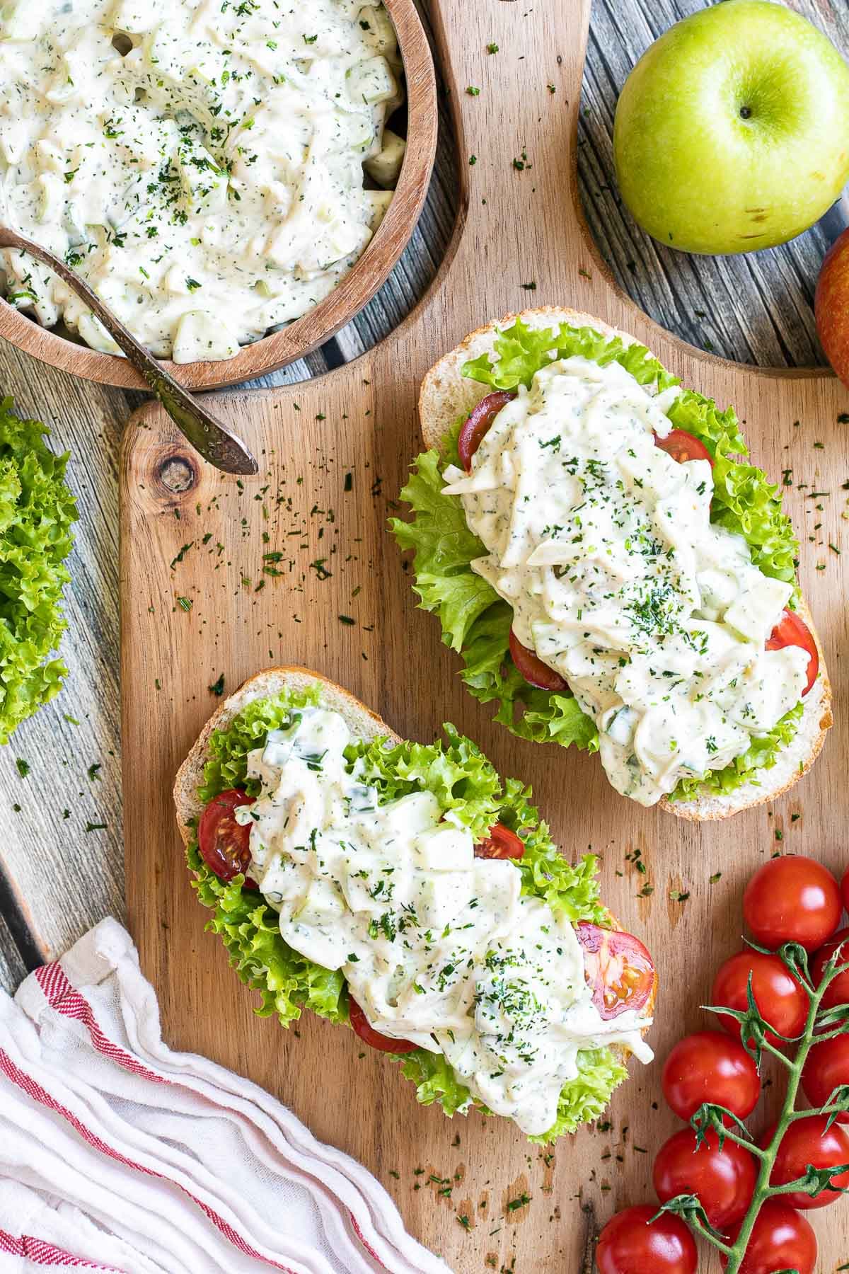 2 small loaves on a wooden board topped with lettuce leaves, cherry tomato pieces and chopped veggies in a white sauce. Leftover juicy white salad is in a wooden bowl.