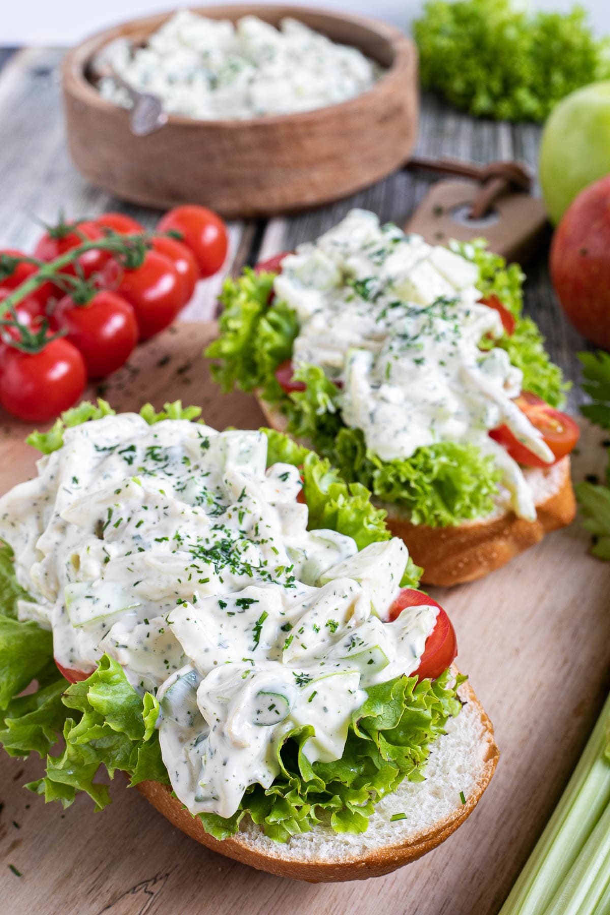 2 small loaves on a wooden board topped with lettuce leaves, cherry tomato pieces and chopped veggies in a white sauce. Leftover juicy white salad is in a wooden bowl.