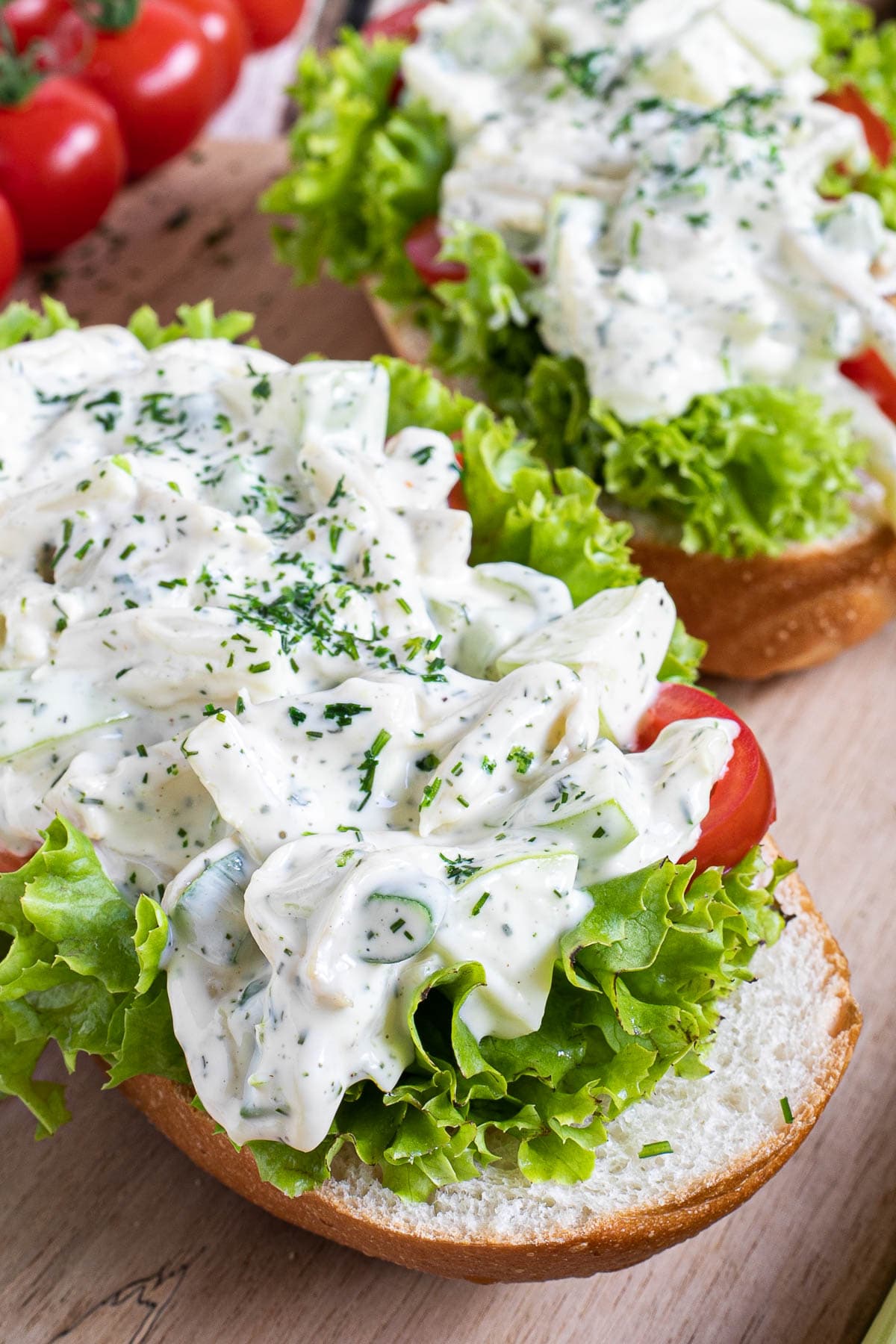 2 small loaves on a wooden board topped with lettuce leaves, cherry tomato pieces and chopped veggies in a white sauce sprinkled with dill and parsley.