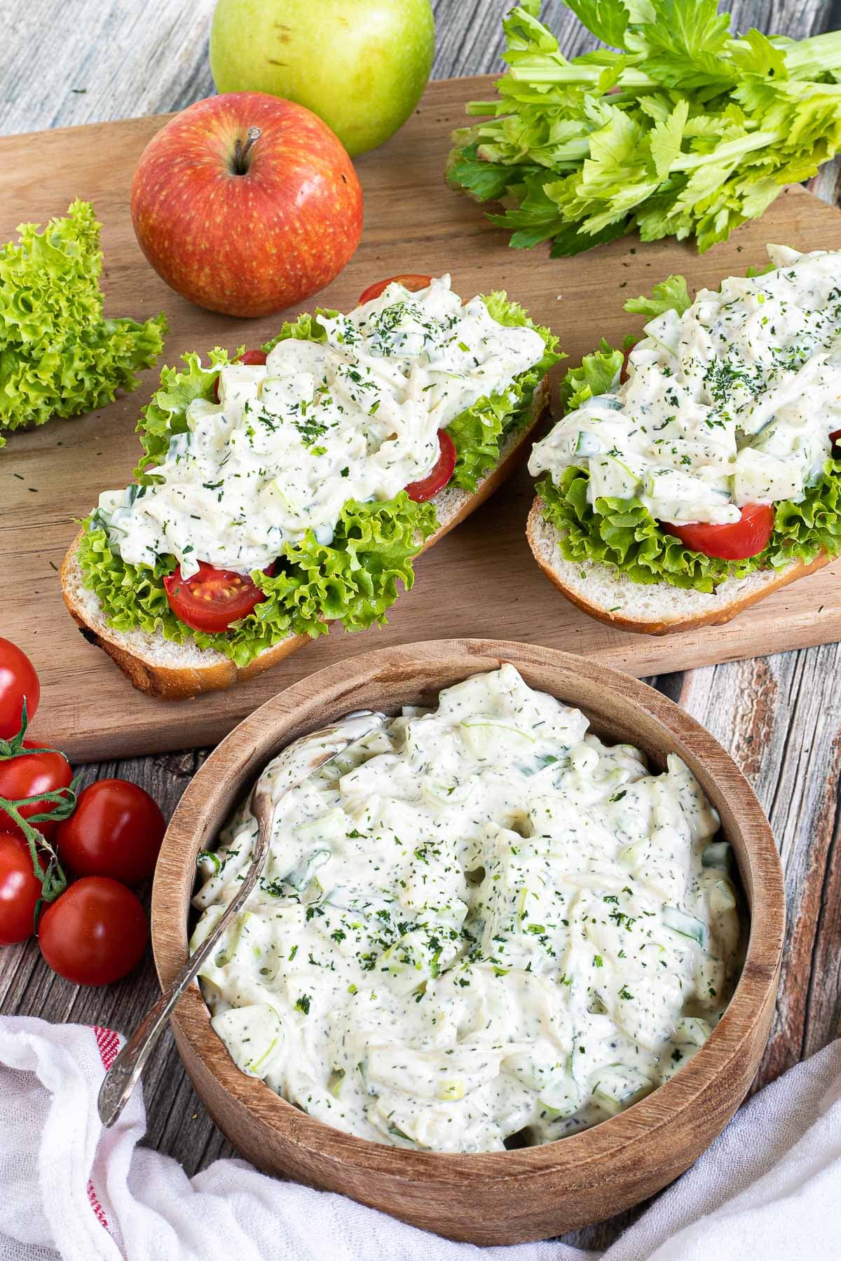 2 small loaves on a wooden board topped with lettuce leaves, cherry tomato pieces and chopped veggies in a white sauce. Leftover juicy white salad is in a wooden bowl.