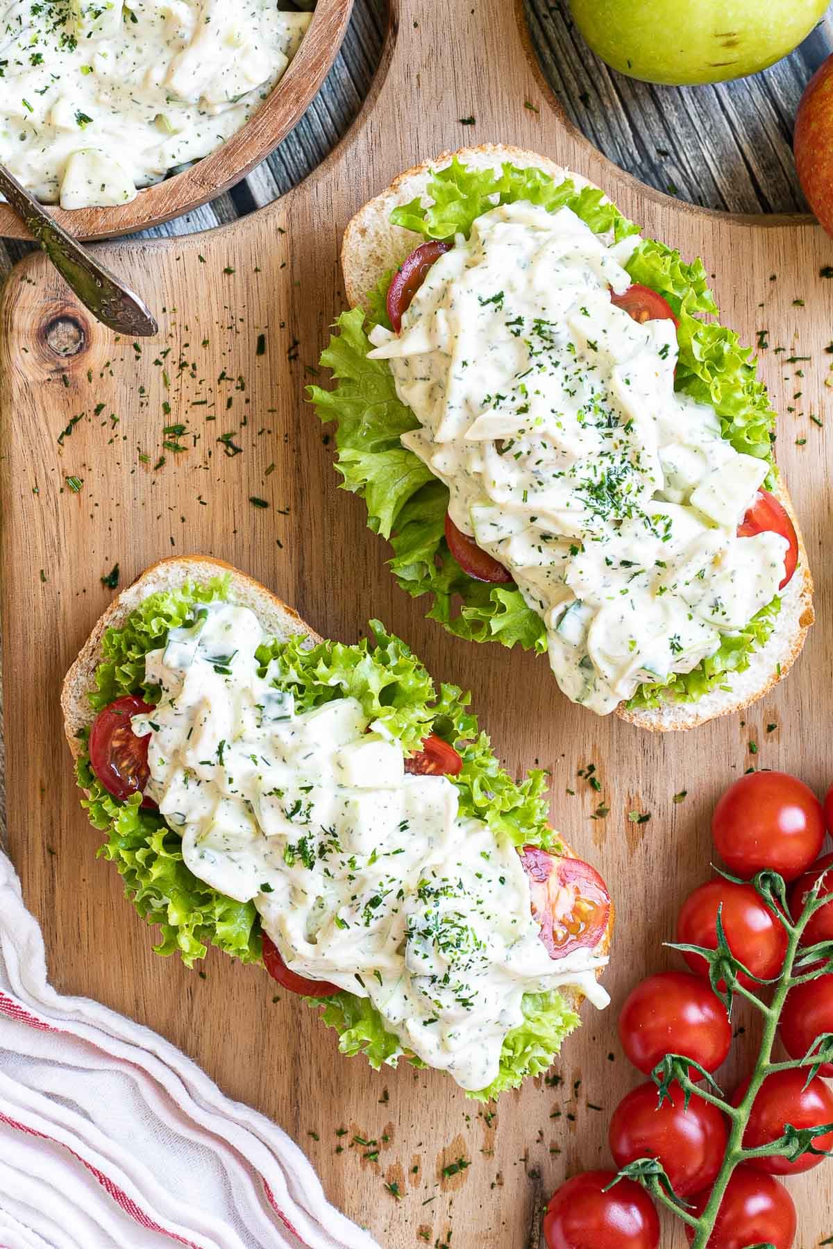 2 small loaves on a wooden board topped with lettuce leaves, cherry tomato pieces and chopped veggies in a white sauce. Leftover juicy white salad is in a wooden bowl.