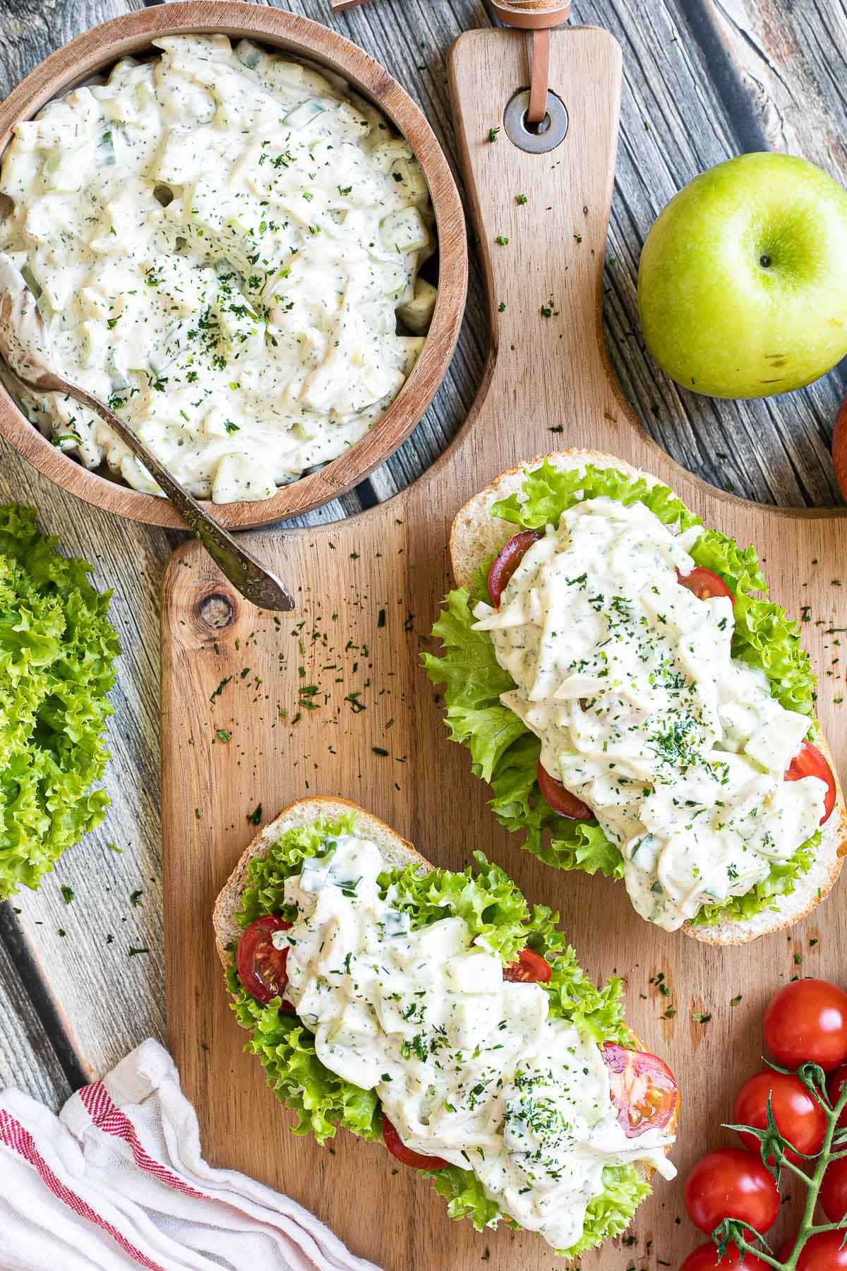 2 small loaves on a wooden board topped with lettuce leaves, cherry tomato pieces and chopped veggies in a white sauce. Leftover juicy white salad is in a wooden bowl.