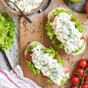 2 small loaves on a wooden board topped with lettuce leaves, cherry tomato pieces and chopped veggies in a white sauce. Leftover juicy white salad is in a wooden bowl.