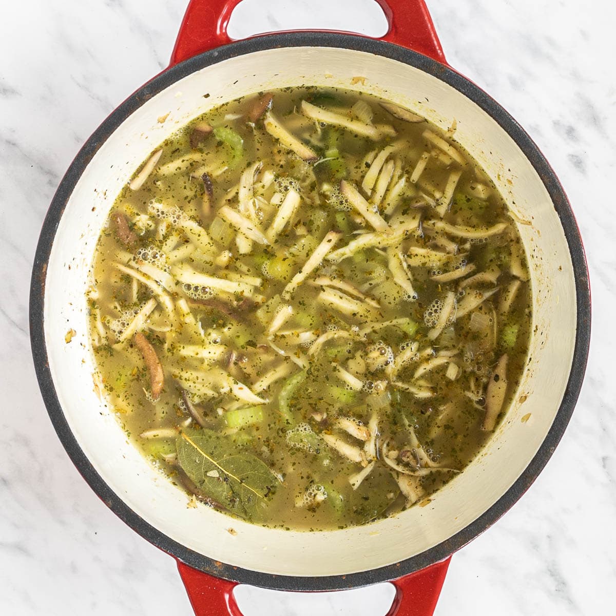 Red Dutch oven with shredded mushrooms, celery slices, chopped onion in a thin light green white soup.