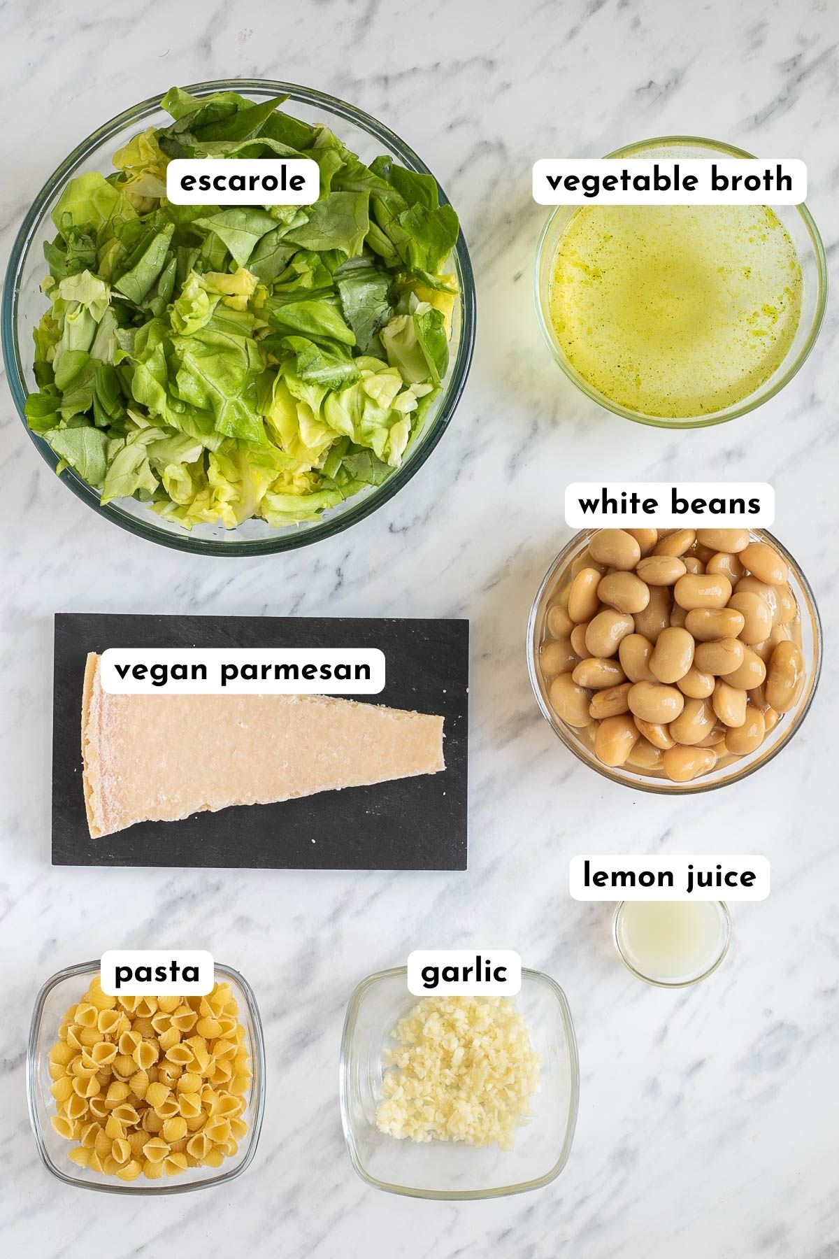 Ingredients of escarole soup in small glass bowls like washed and chopped escarole, vegetable broth, white beans, small shell pasta, chopped garlic, lemon juice, and a block of parmesan cheese