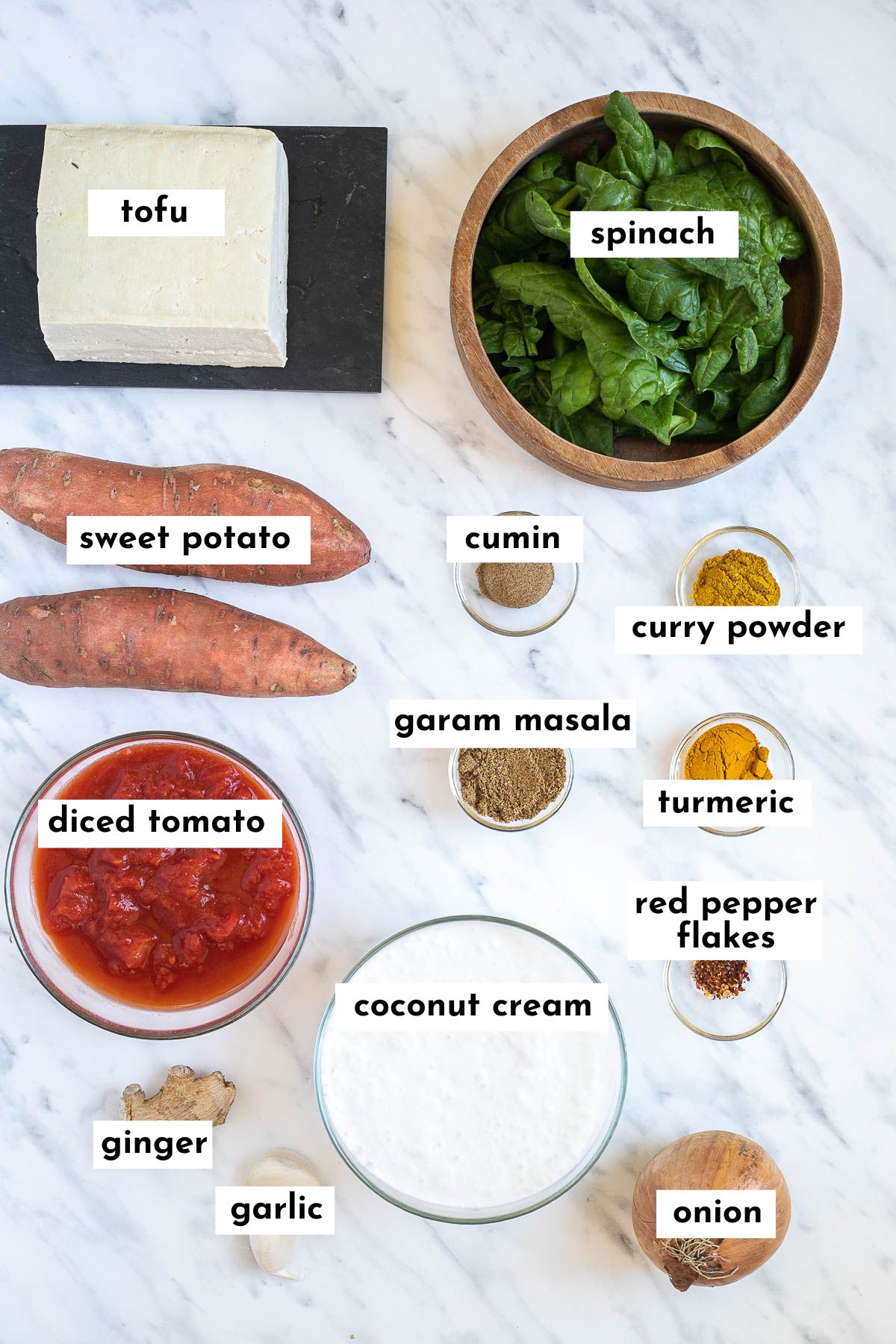 Ingredients of sweet potato tofu curry in small glass bowls or on a marble surface like tofu block, spinach leaves, sweet potatoes, diced tomatoes, coconut cream, onion and various spices. 
