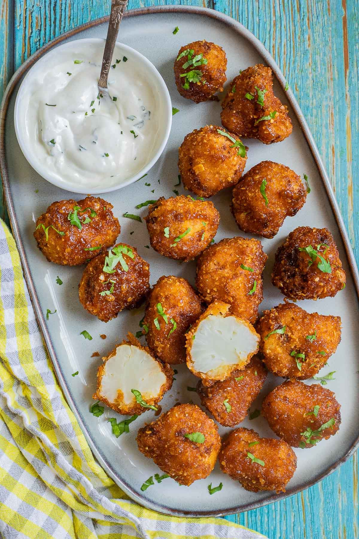 Breaded cauliflower florets in a light blue plate sprinkled with chopped green herbs and served with a small bowl of white sauce. One floret is cut in half to show the white insides.