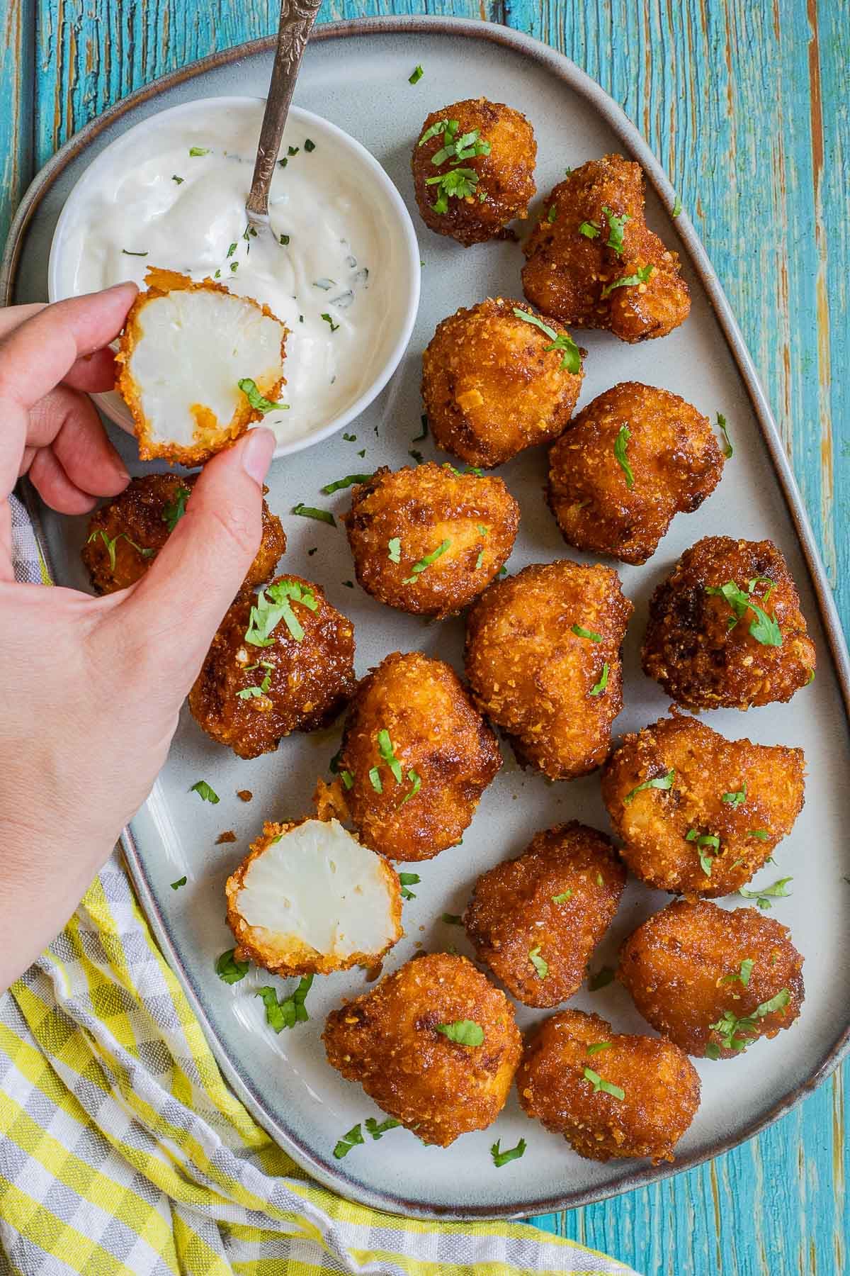 Breaded cauliflower florets in a light blue plate sprinkled with chopped green herbs and served with a small bowl of white sauce. One floret is cut in half to show the white insides. A hand is holding one half and about to dip it.