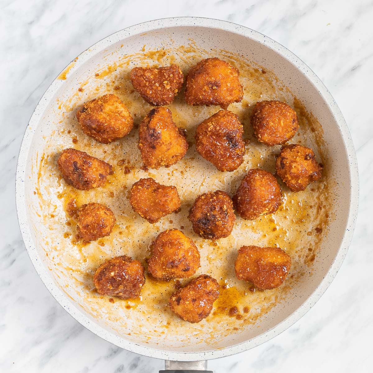 Breaded cauliflower florets are thinly covered in a light orange sauce in a white frying pan