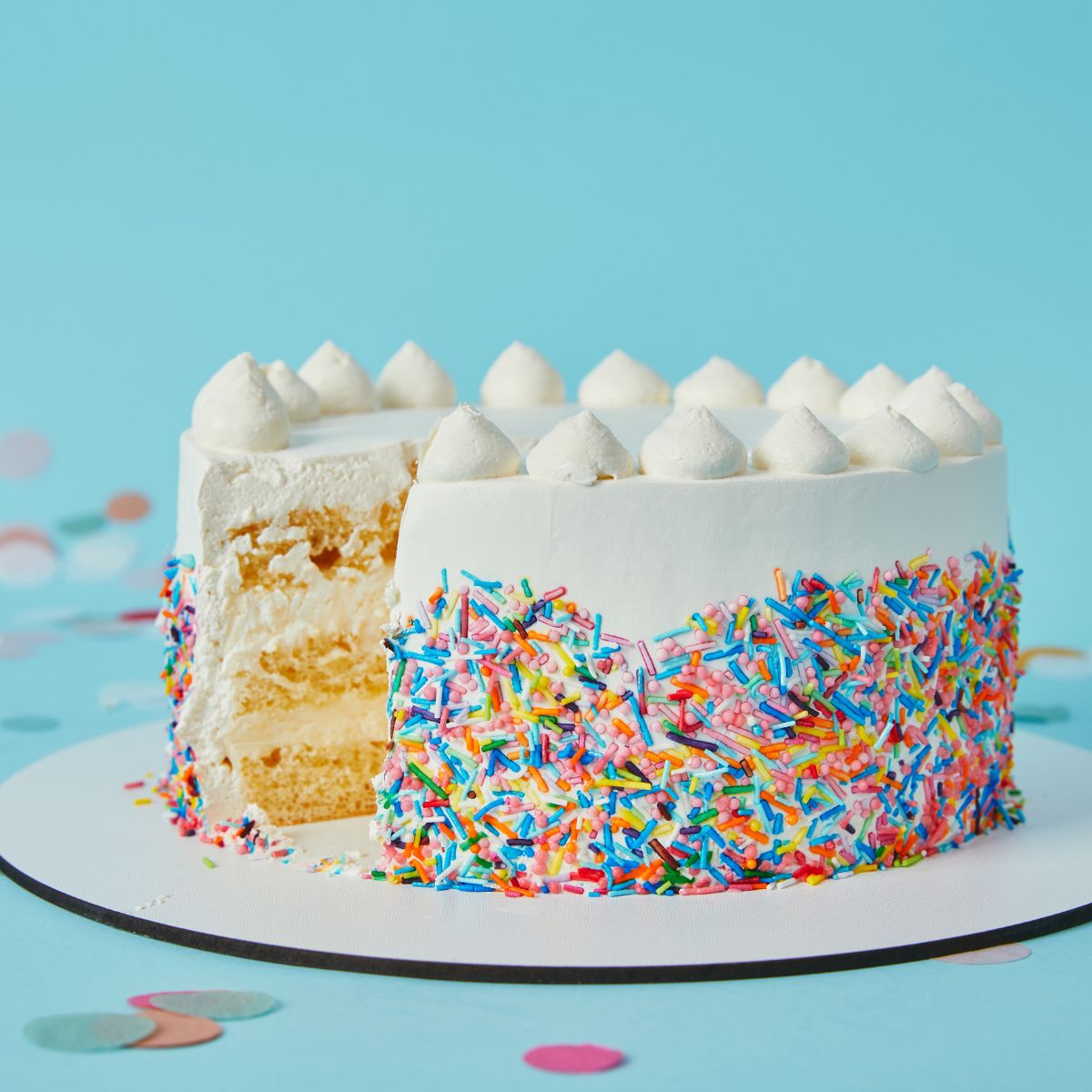 A white vanilla cake decorated with colorful sprinkles and some white frosting on top is placed in a white disk on a light blue surface. 