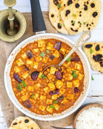 A white frying pan with tofu cubes, chickpeas and large red onion pieces mixed with a red-orange sauce and topped with freshly chopped herbs. Several pieces of flatbread and a wooden bowl of rice are next to it.