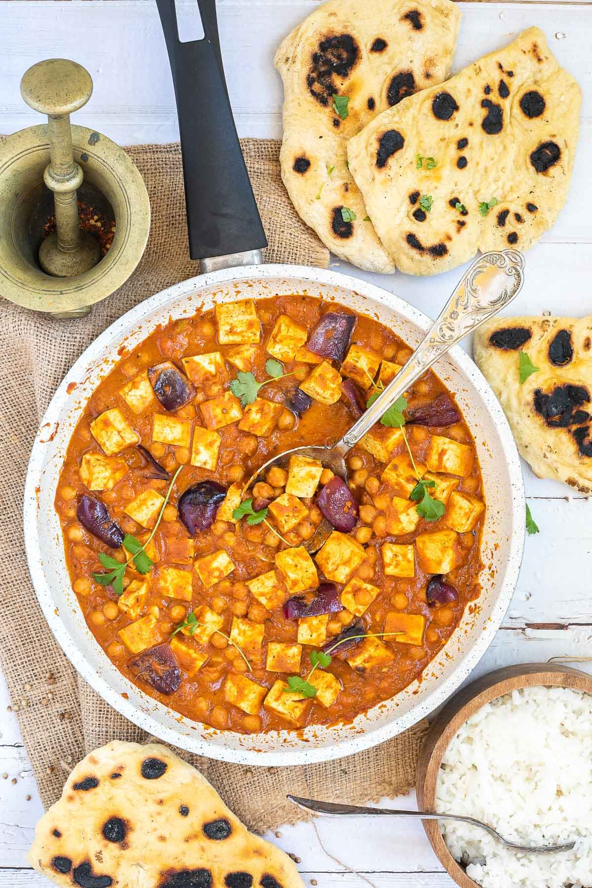 A white frying pan with tofu cubes, chickpeas and large red onion pieces mixed with a red-orange sauce and topped with freshly chopped herbs. Several pieces of flatbread and a wooden bowl of rice are next to it. 