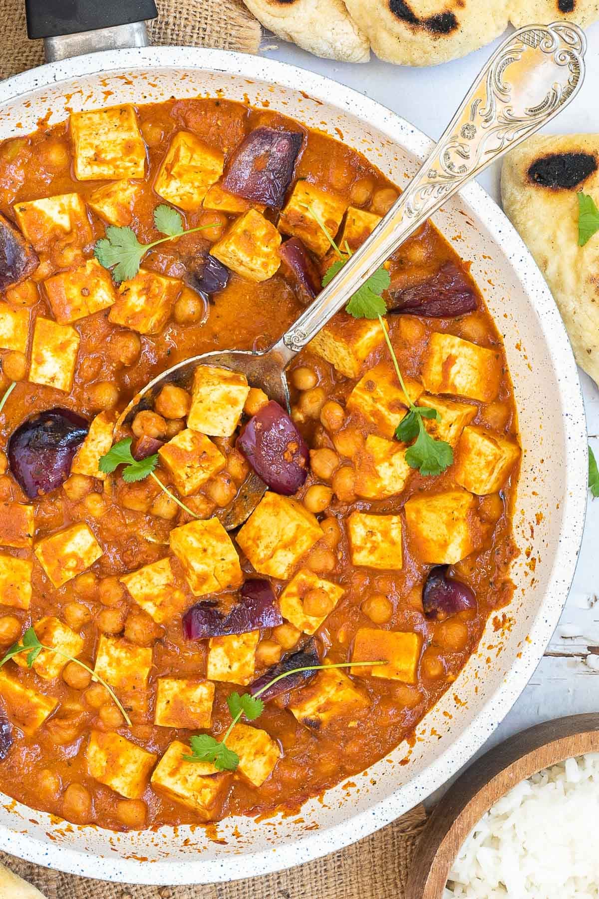 A white frying pan with tofu cubes, chickpeas and large red onion pieces mixed with a red-orange sauce and topped with freshly chopped herbs.