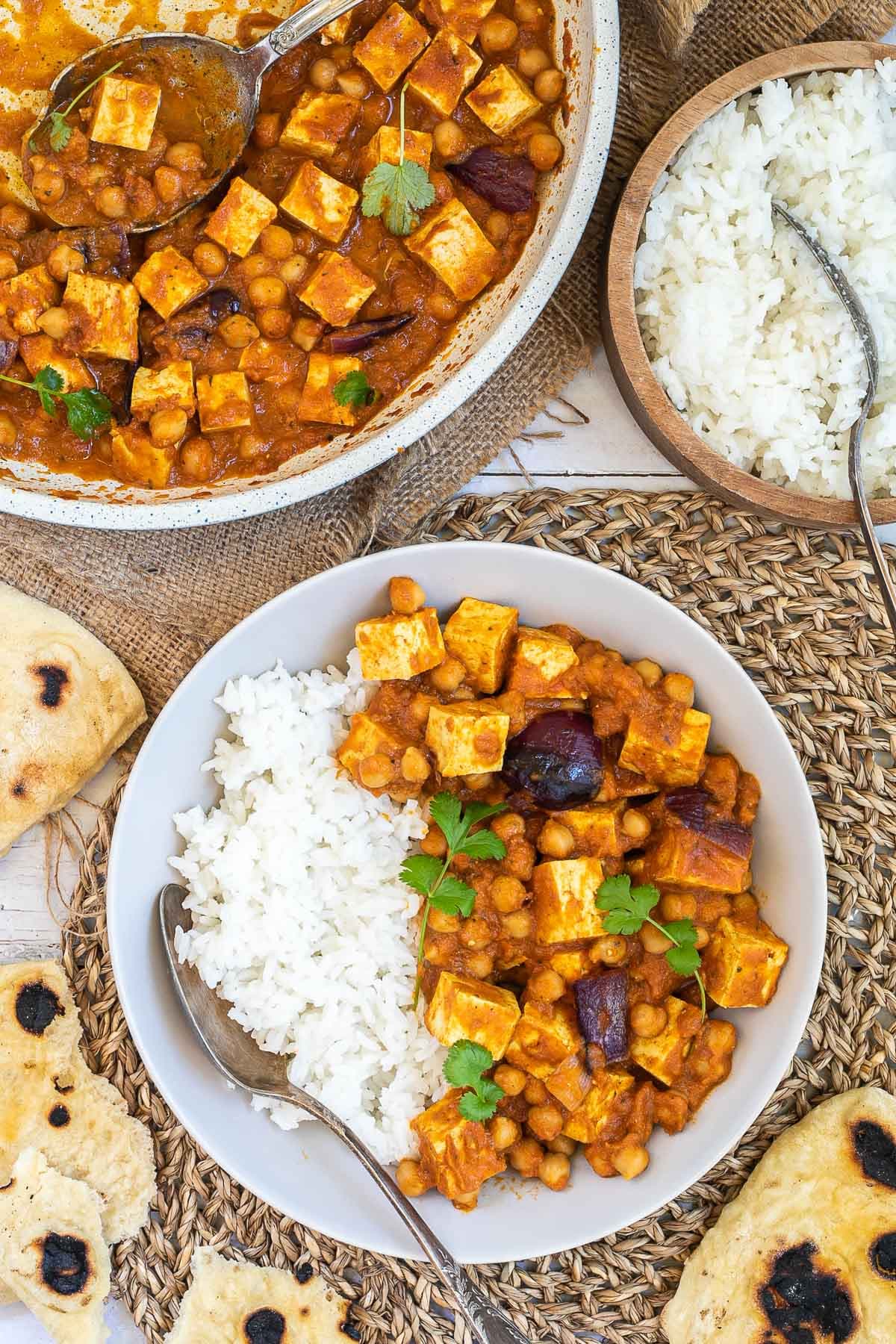 A white bowl with tofu cubes, chickpeas, and large red onion pieces mixed with a red-orange sauce served next to white rice. Leftovers in a white frying pan, several pieces of flatbread and a wooden bowl of rice are next to it.