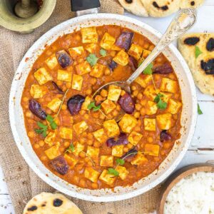 A white frying pan with tofu cubes, chickpeas and large red onion pieces mixed with a red-orange sauce and topped with freshly chopped herbs. Several pieces of flatbread and a wooden bowl of rice are next to it.