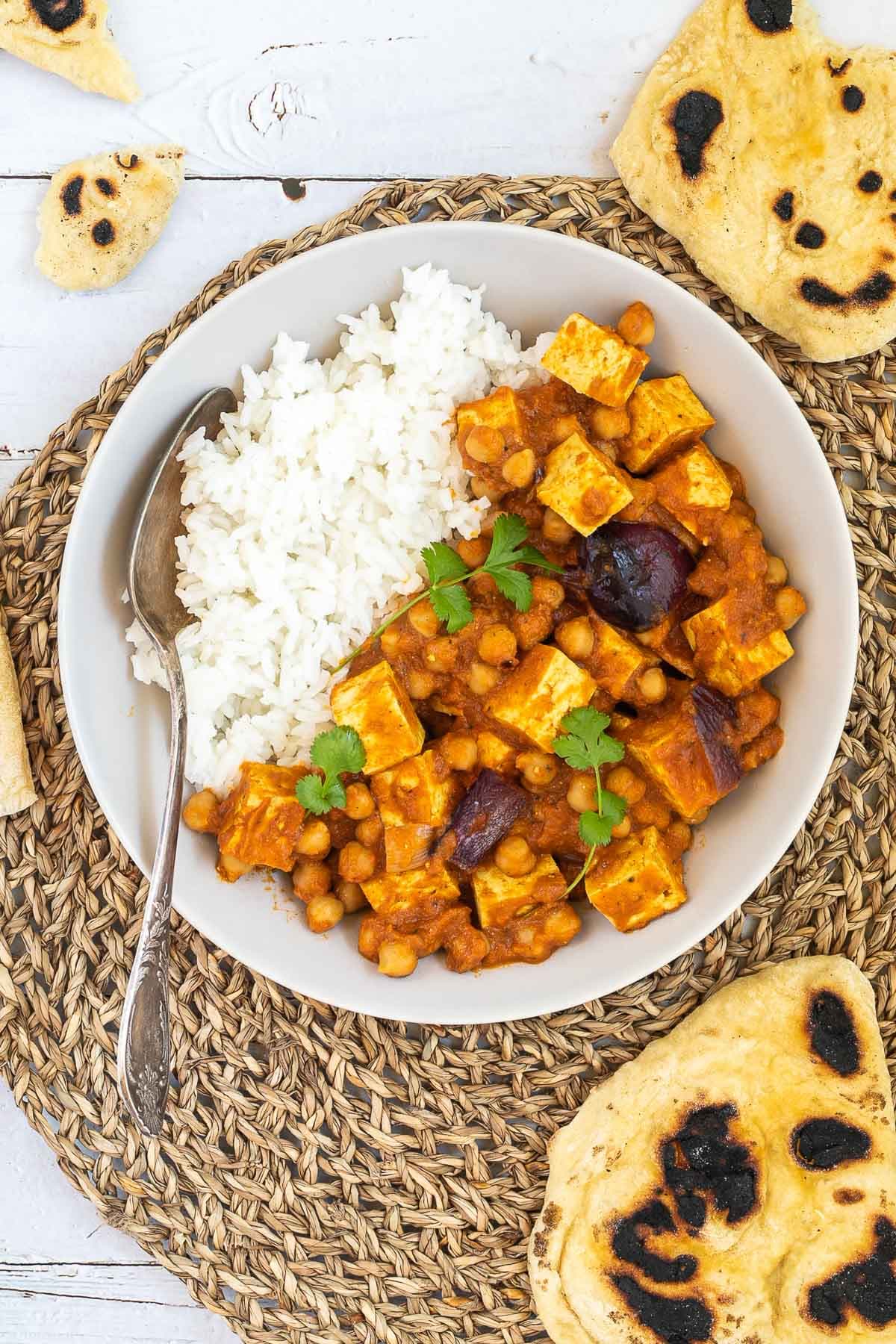 A white bowl with tofu cubes, chickpeas, and large red onion pieces mixed with a red-orange sauce served next to white rice. Several pieces of flatbread are next to it.