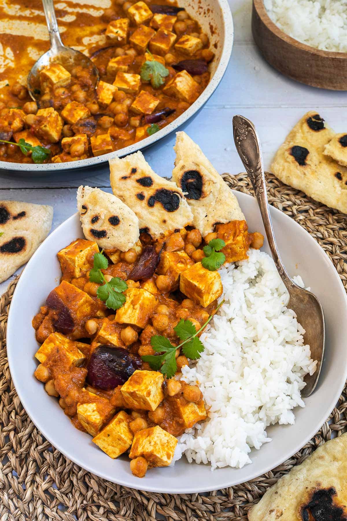 A white bowl with tofu cubes, chickpeas, and large red onion pieces mixed with a red-orange sauce served next to white rice and pieces of flatbread. Leftovers in a white frying pan, several pieces of flatbread and a wooden bowl of rice are next to it.