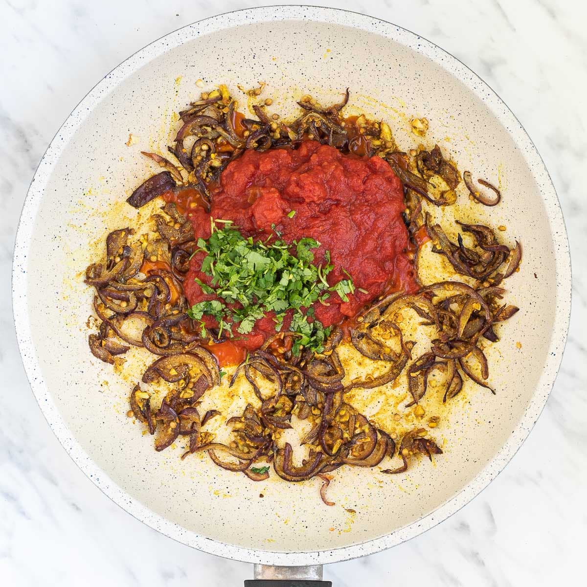 White frying pan with brown sauteed onion slices, tomato sauce and chopped green herbs