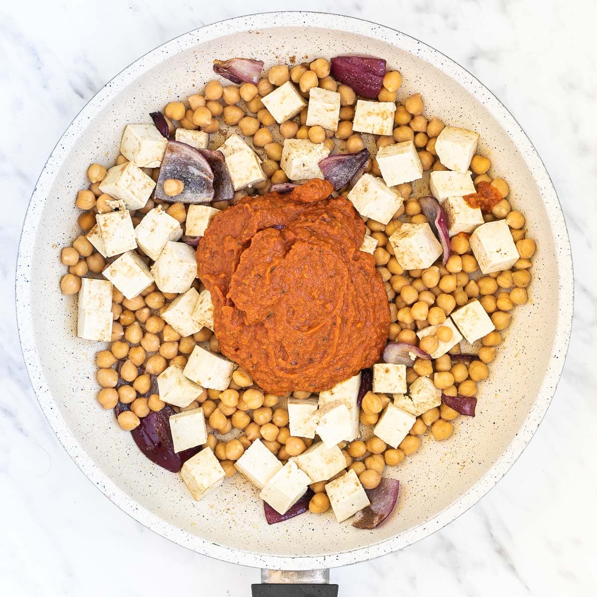 Red onion pieces, tofu cubes, and chickpeas with a thick red sauce on top in a white frying pan.