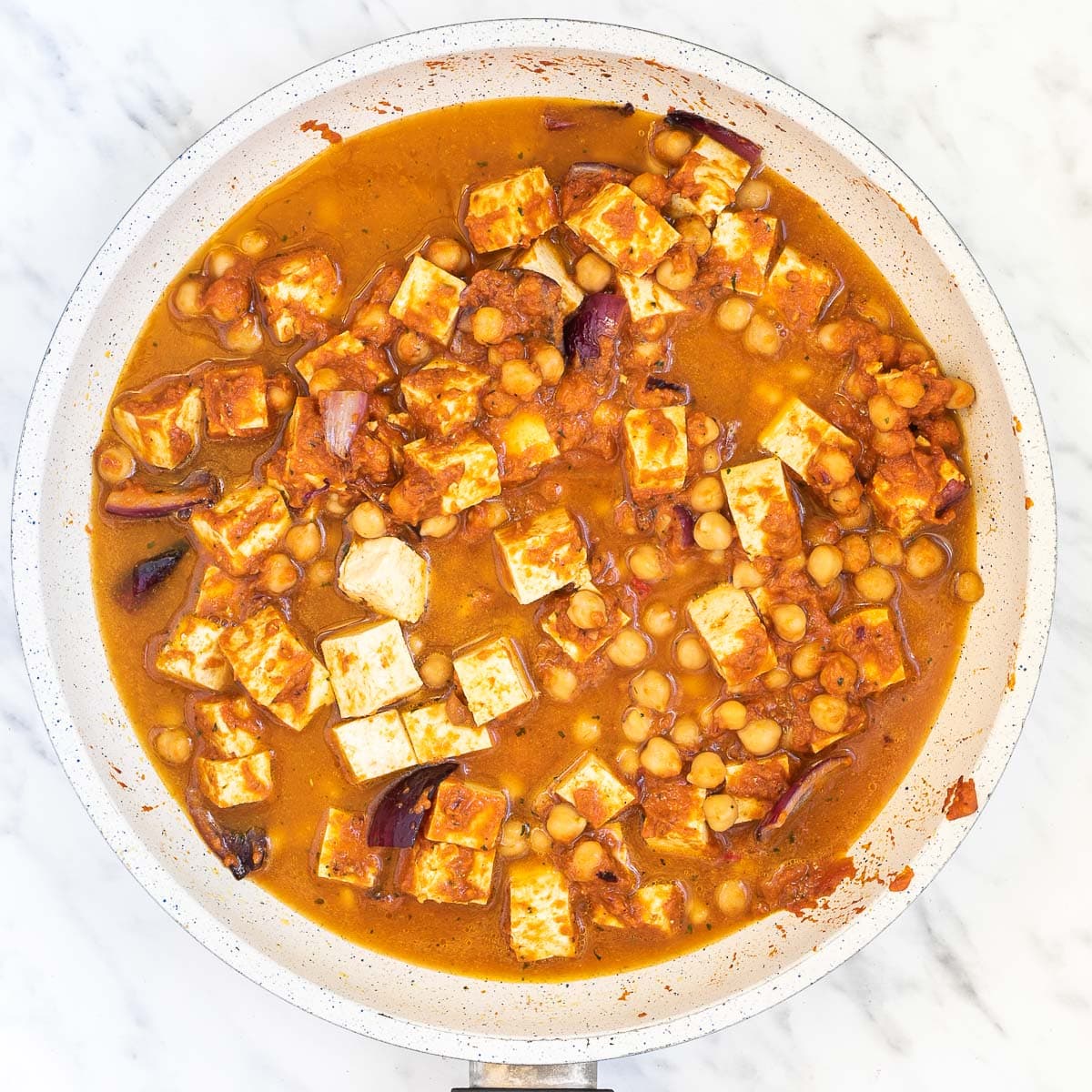 Red onion pieces, tofu cubes, and chickpeas mixed with a thick red orange sauce in a white frying pan.