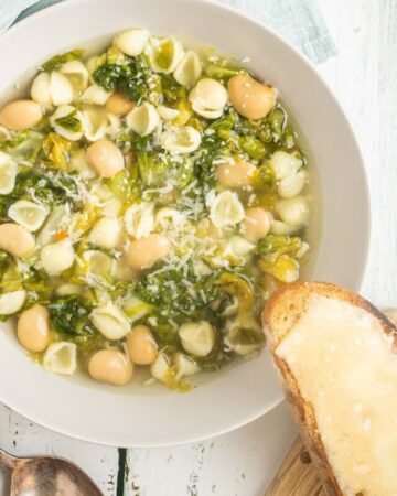 A white bowl of thin soup with white beans, chopped green escarole, small shell pasta, topped with grated cheese. A cheesy, toasted bread is placed on the side of the bowl.